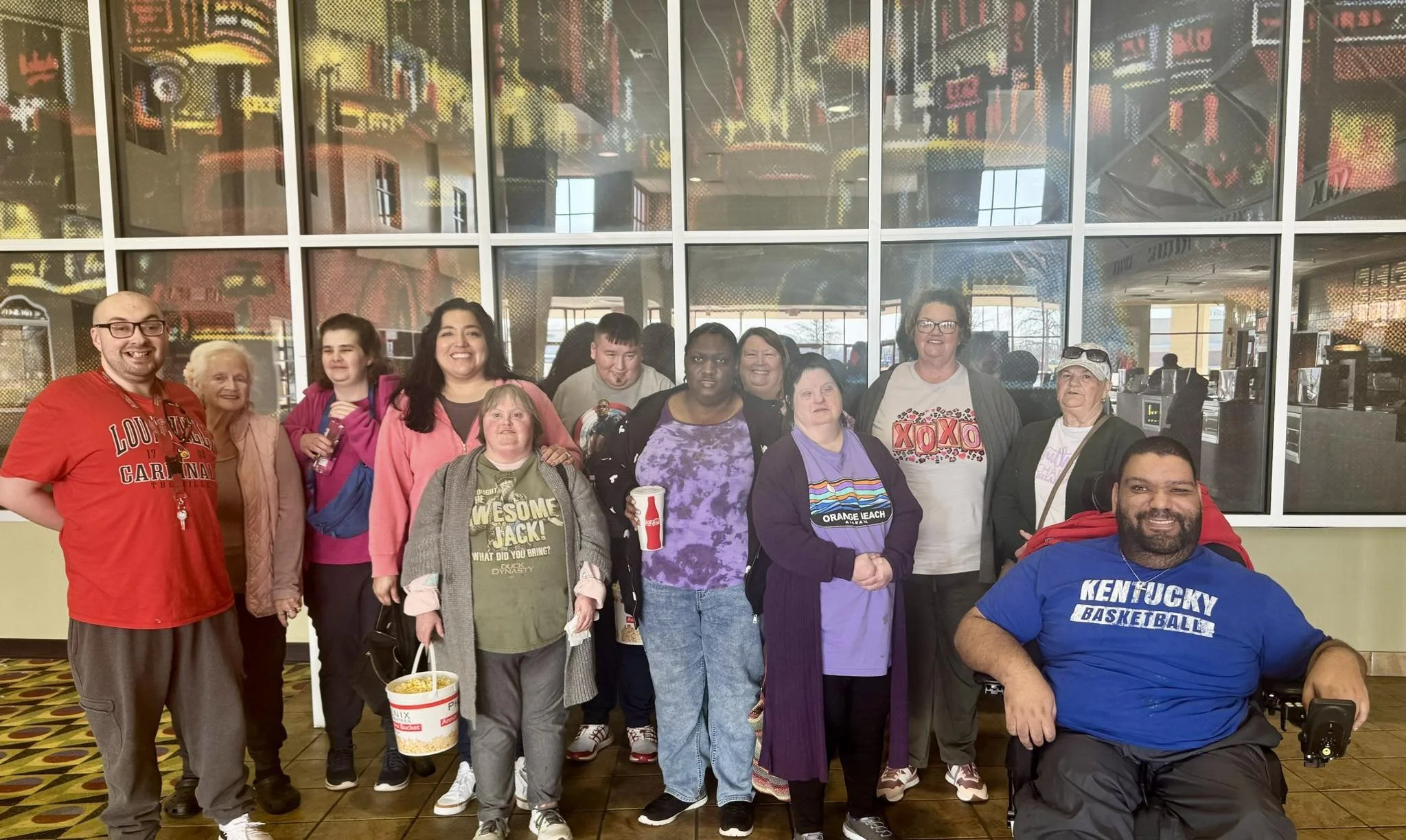 Group of diverse people smiling and posing for a photo in an indoor location with large glass windows and a colorful, abstract mural in the background.