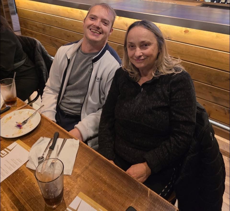 Two people sitting at a restaurant table with empty plates and drinks, smiling for the camera. The background features wooden paneling.
