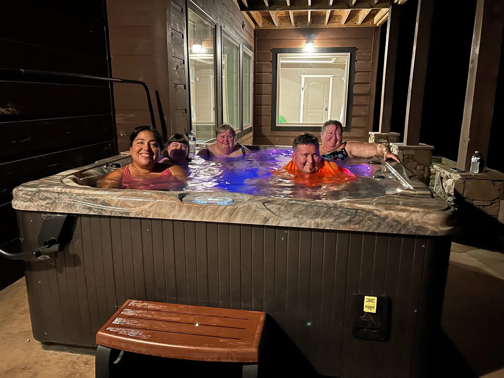 A group of five people in a hot tub at night, smiling and enjoying each other's company, with a wooden deck and a house exterior in the background.