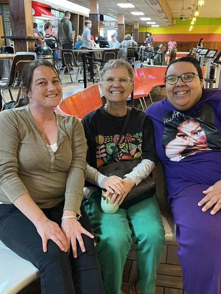 Three women sitting together in a food court at a mall or shopping center, smiling at the camera. The background shows other people seated at tables and standing near counters.