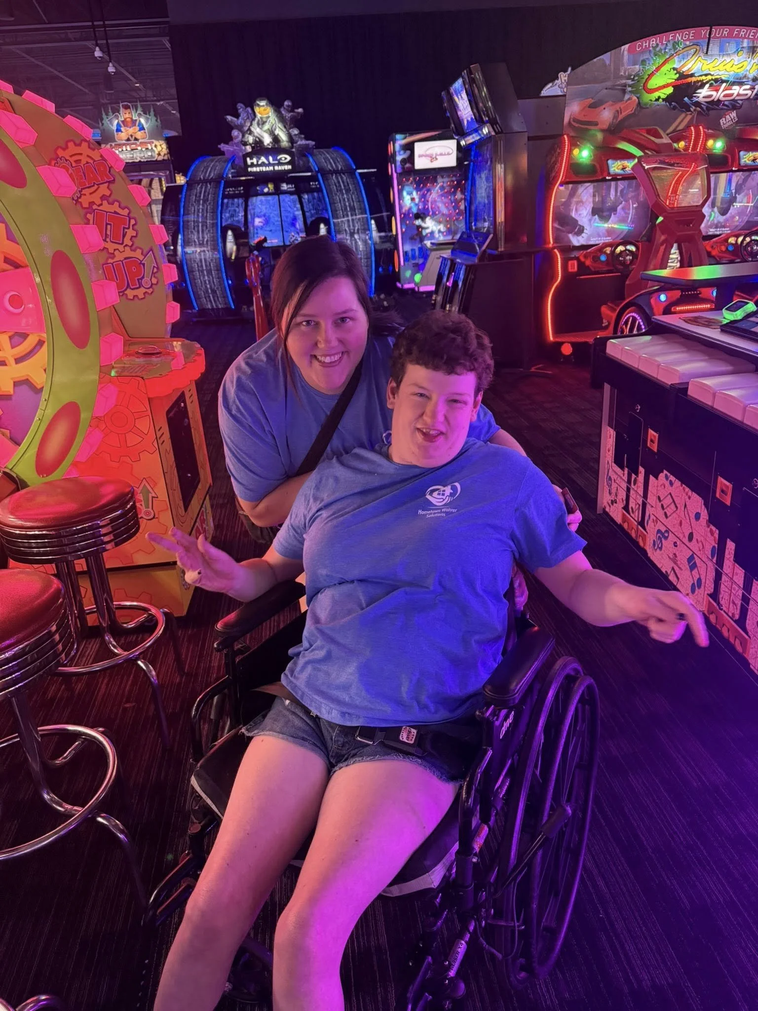 Two people smiling and posing in an arcade, one sitting in a wheelchair making peace signs, with colorful arcade games and neon lights in the background.