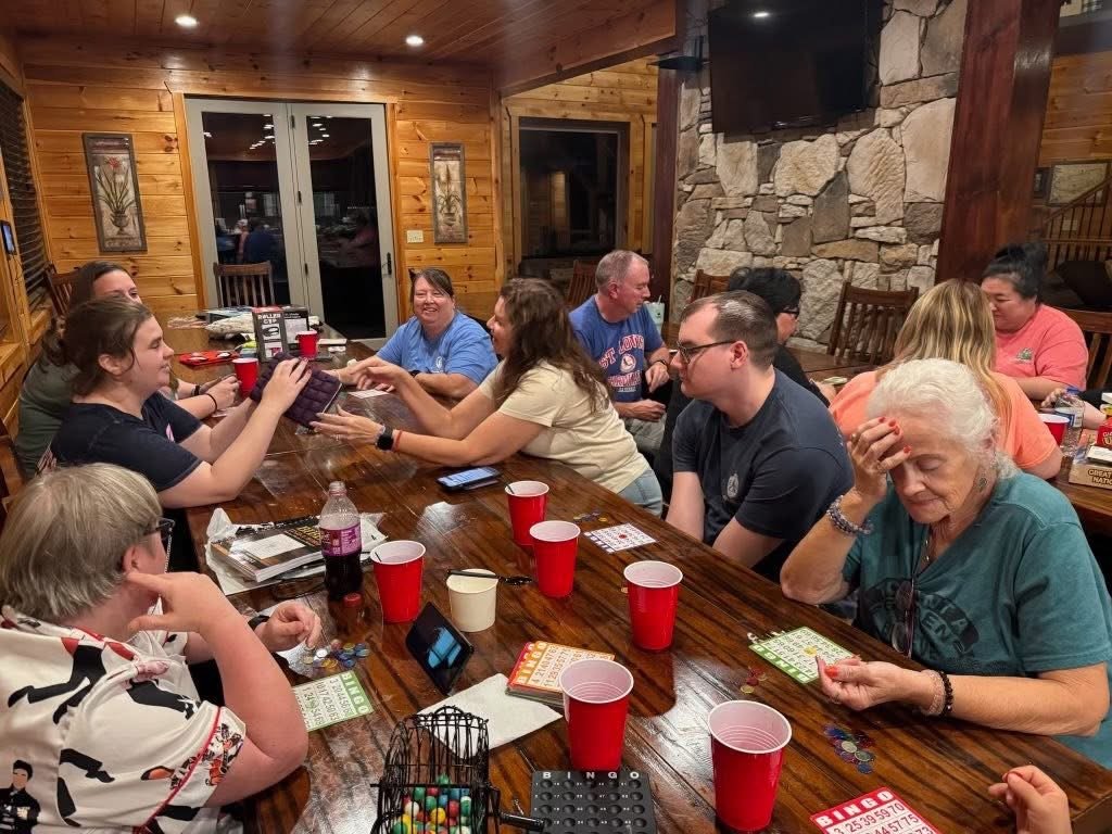Group of people sitting around a wooden table in a cozy cabin with wood-paneled walls and a stone fireplace, engaged in a group activity or game, with some smiling and talking.