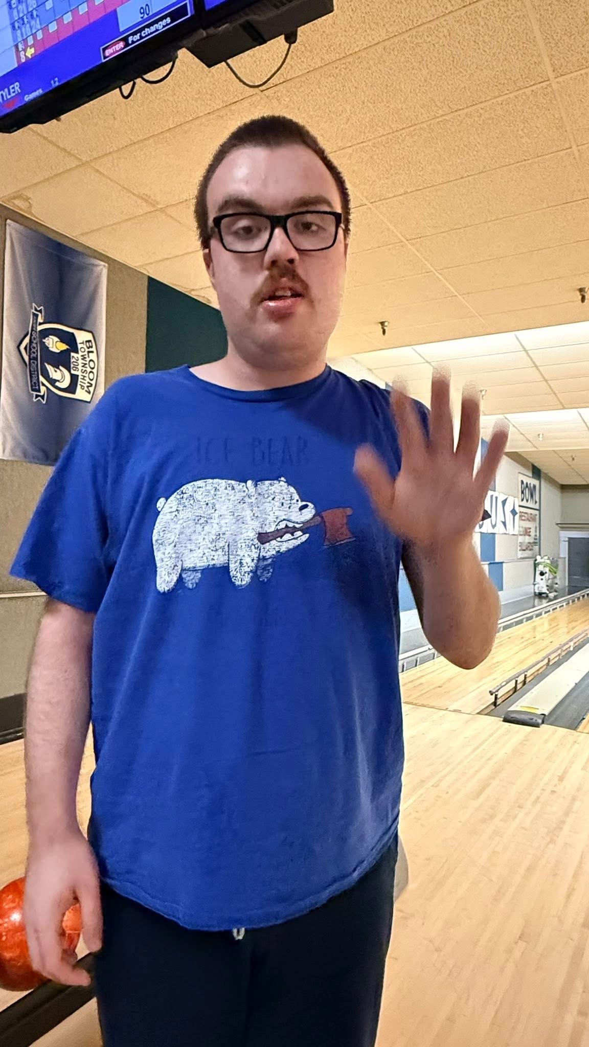 Young man with glasses and short hair waving, standing in a bowling alley, holding an orange bowling ball, wearing a blue t-shirt with a bear graphic.