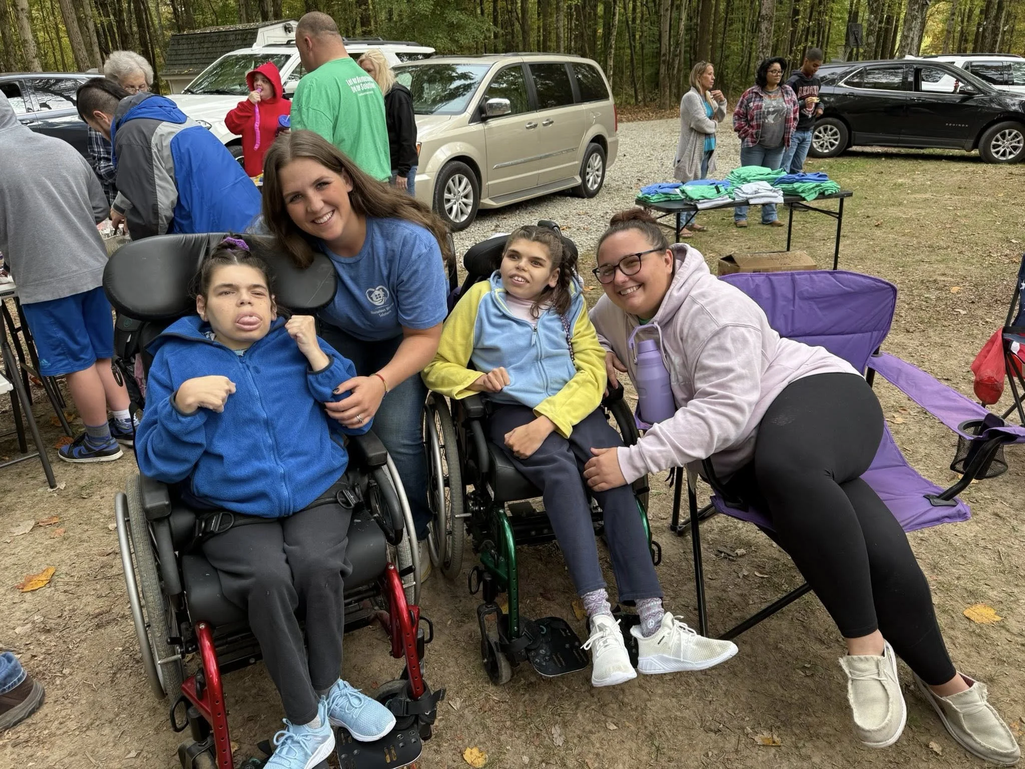 A group of people, including children in wheelchairs, gathers outdoors at a park or event, smiling and enjoying the day with trees and parked cars in the background.