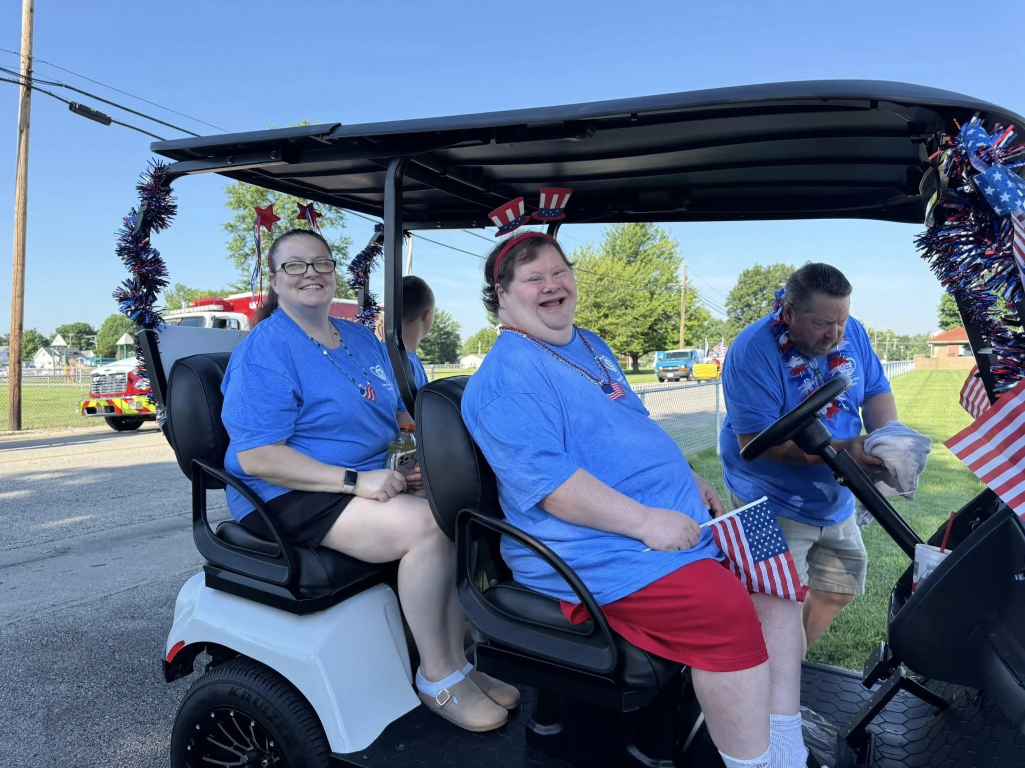 Three people dressed in patriotic blue shirts with American flags riding in a decorated golf cart during a parade, with one person wearing a headband with red, white, and blue stars, and another with red, white, and blue pinwheel headbands.