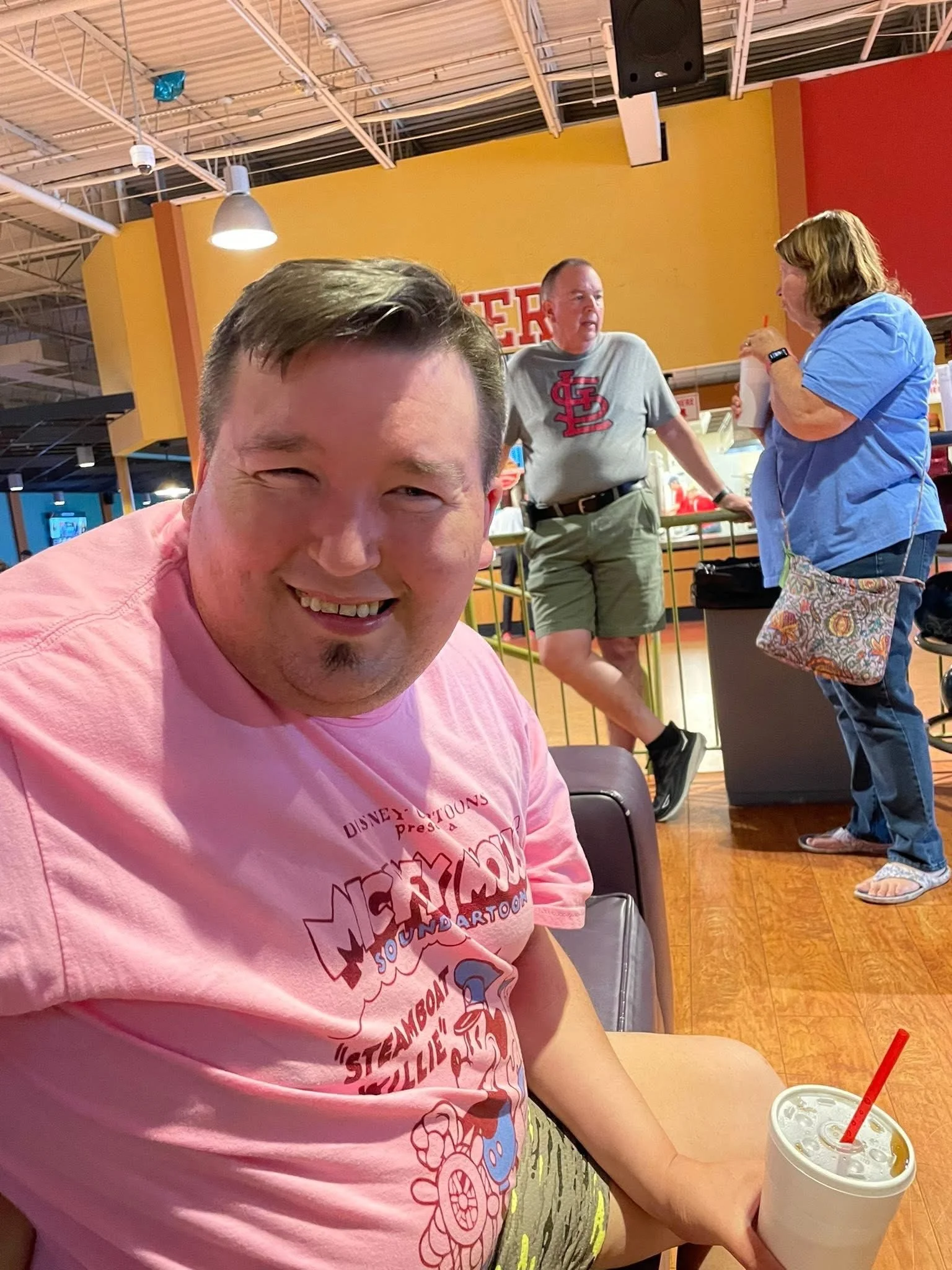 A smiling man wearing a pink T-shirt sitting inside a restaurant or cafe, holding a cup with a straw. In the background, two people are talking, one wearing a gray T-shirt with a red logo and shorts, the other wearing a blue shirt and jeans, holding 