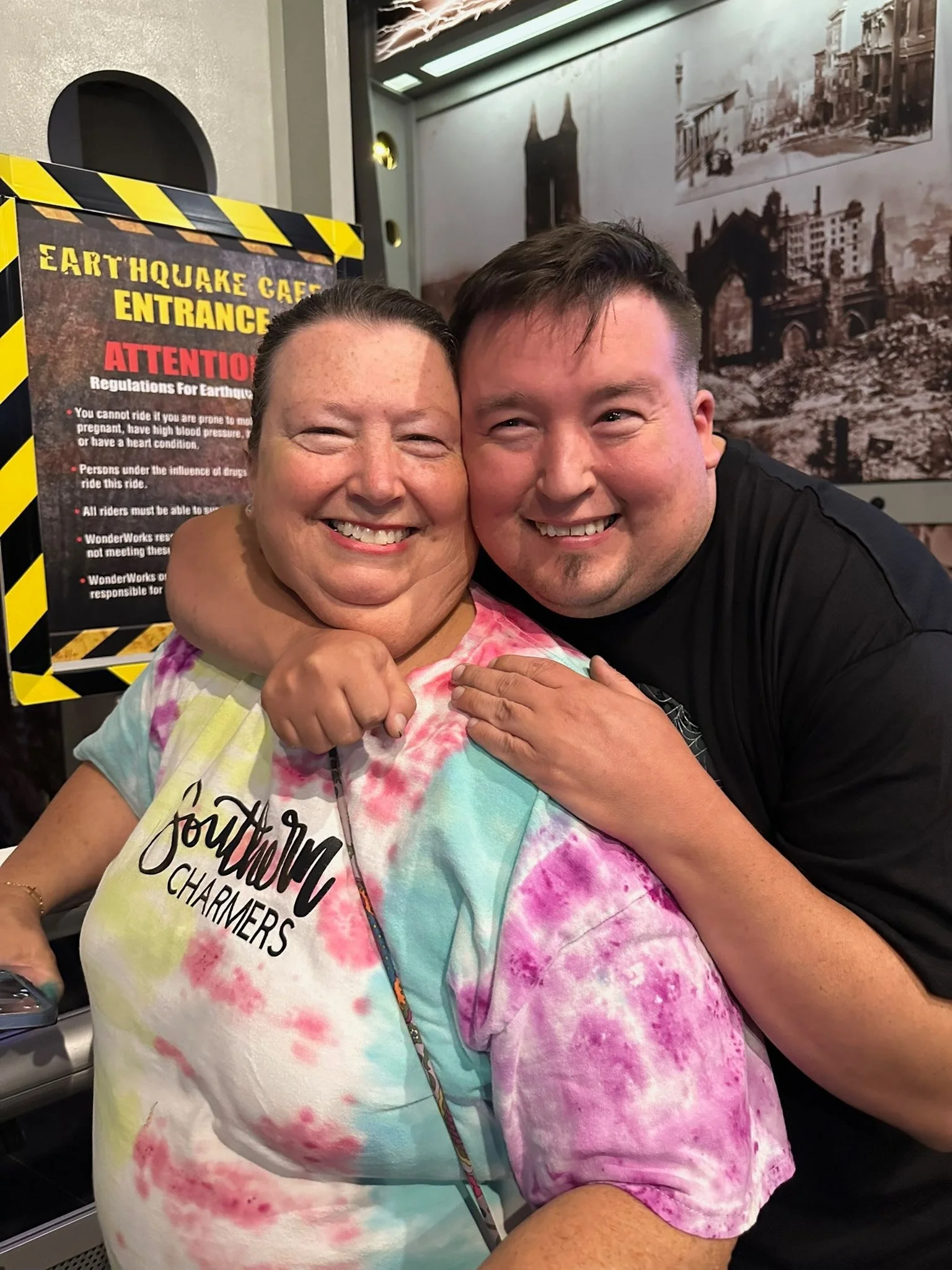 A smiling woman with short dark hair in a colorful tie-dye shirt and a smiling young man with short dark hair in a black shirt hug each other in a friendly pose.