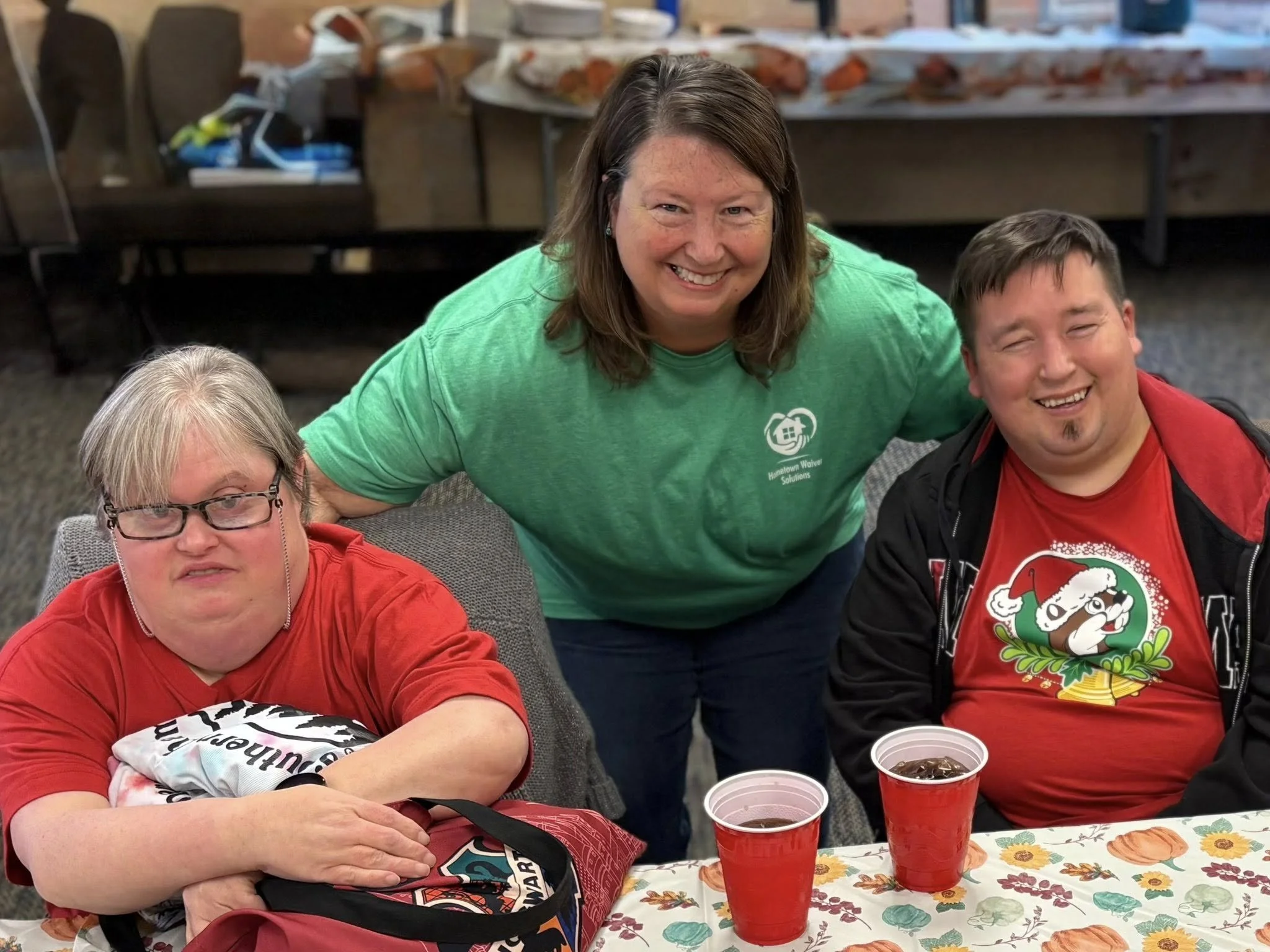 Three people smiling at a table decorated with a fall-themed tablecloth, two sitting and one standing behind them. The woman in the middle is wearing a green shirt, and the two seated individuals are wearing red holiday-themed shirts. There are two r