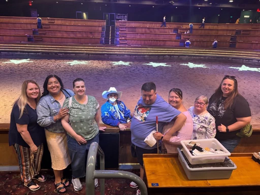 Group of eight people standing in front of a rodeo arena, smiling, with wooden seating and a dirt rodeo ring in the background.