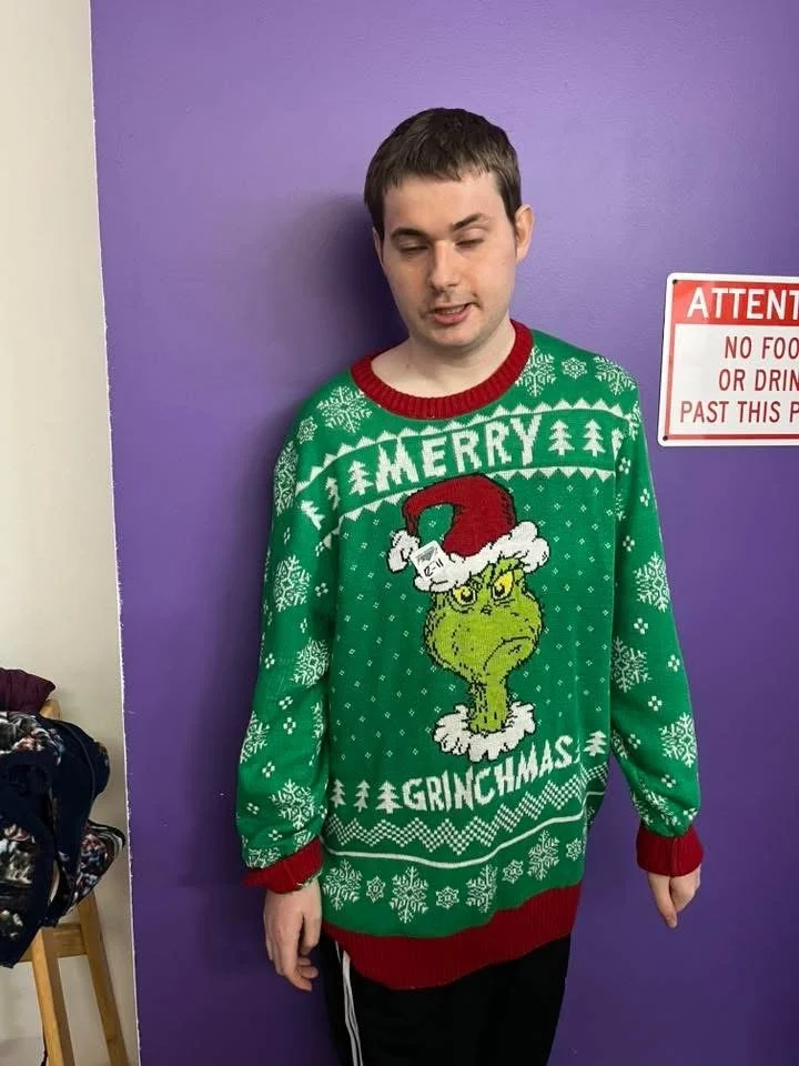 A young man wearing a green Christmas sweater featuring the Grinch with a Santa hat and the words 'Merry Grinchmas' in front of a purple wall.