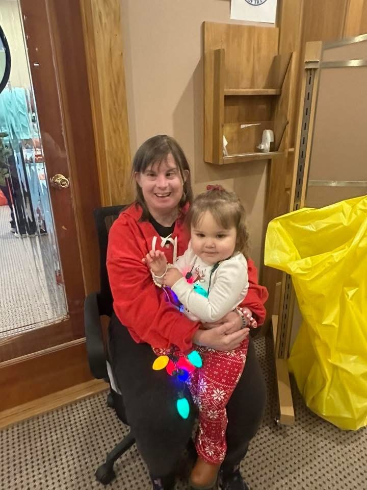 A woman with short brown hair wearing a red hoodie sits in a black office chair, smiling, while holding a young girl with brown hair and a bow, dressed in holiday red patterned pajamas. The girl has colorful holiday lights wrapped around her waist.