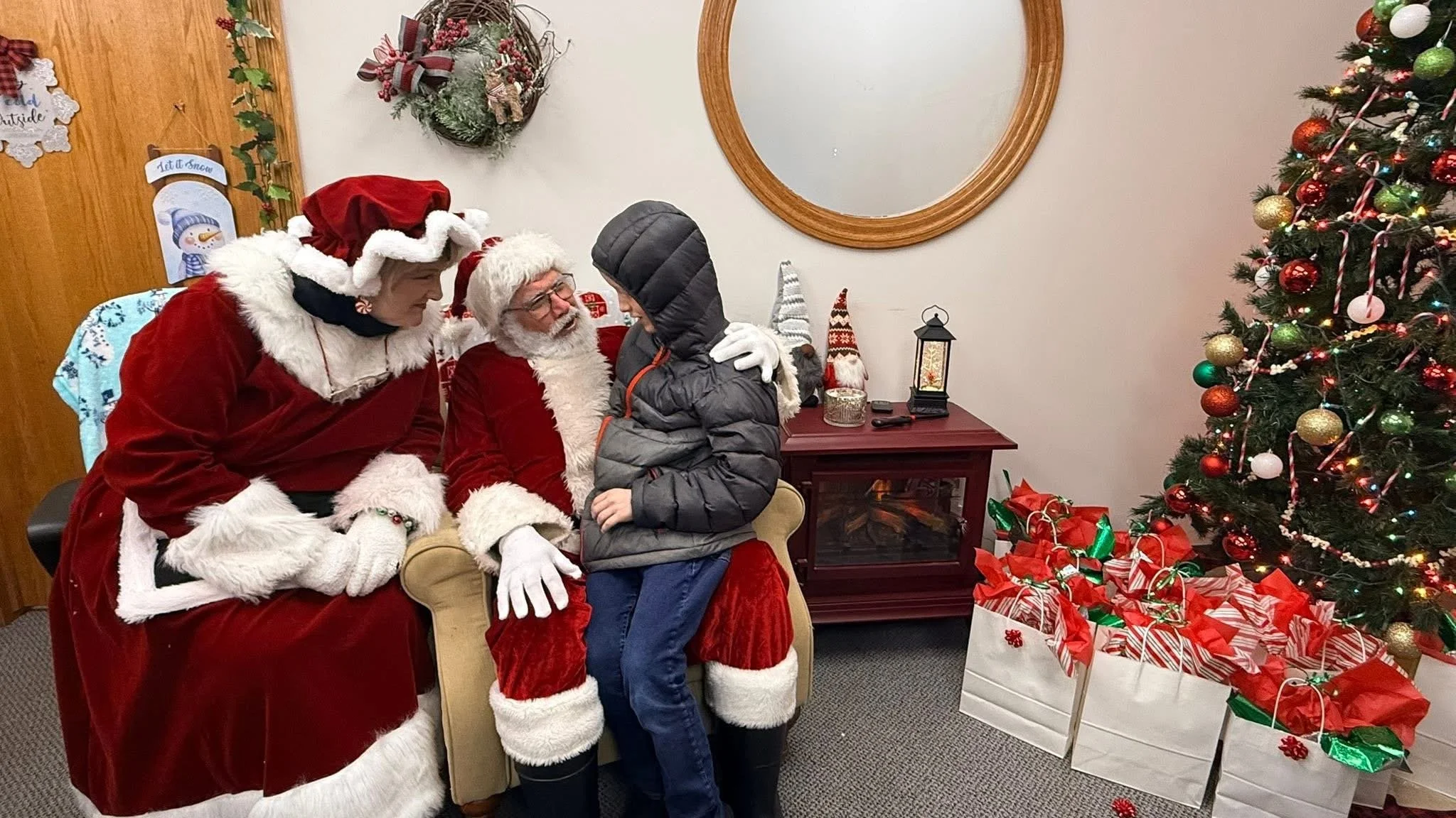 A child sitting on Santa Claus's lap talking to him, with a woman dressed as Mrs. Claus sitting next to Santa, in a room decorated for Christmas with a Christmas tree, presents, and holiday decorations.