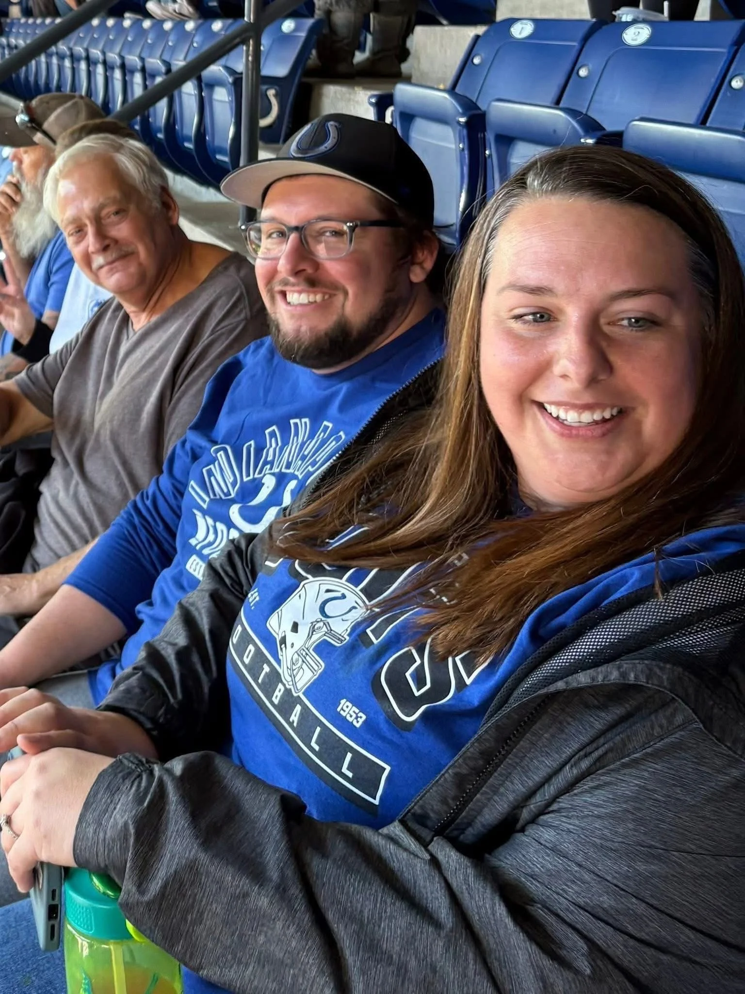 Group of people sitting in stadium seats, smiling for a selfie. The woman in the foreground has long brown hair, is wearing a black and blue sports shirt, and is holding a green water bottle. The man next to her has glasses, a beard, and is wearing a