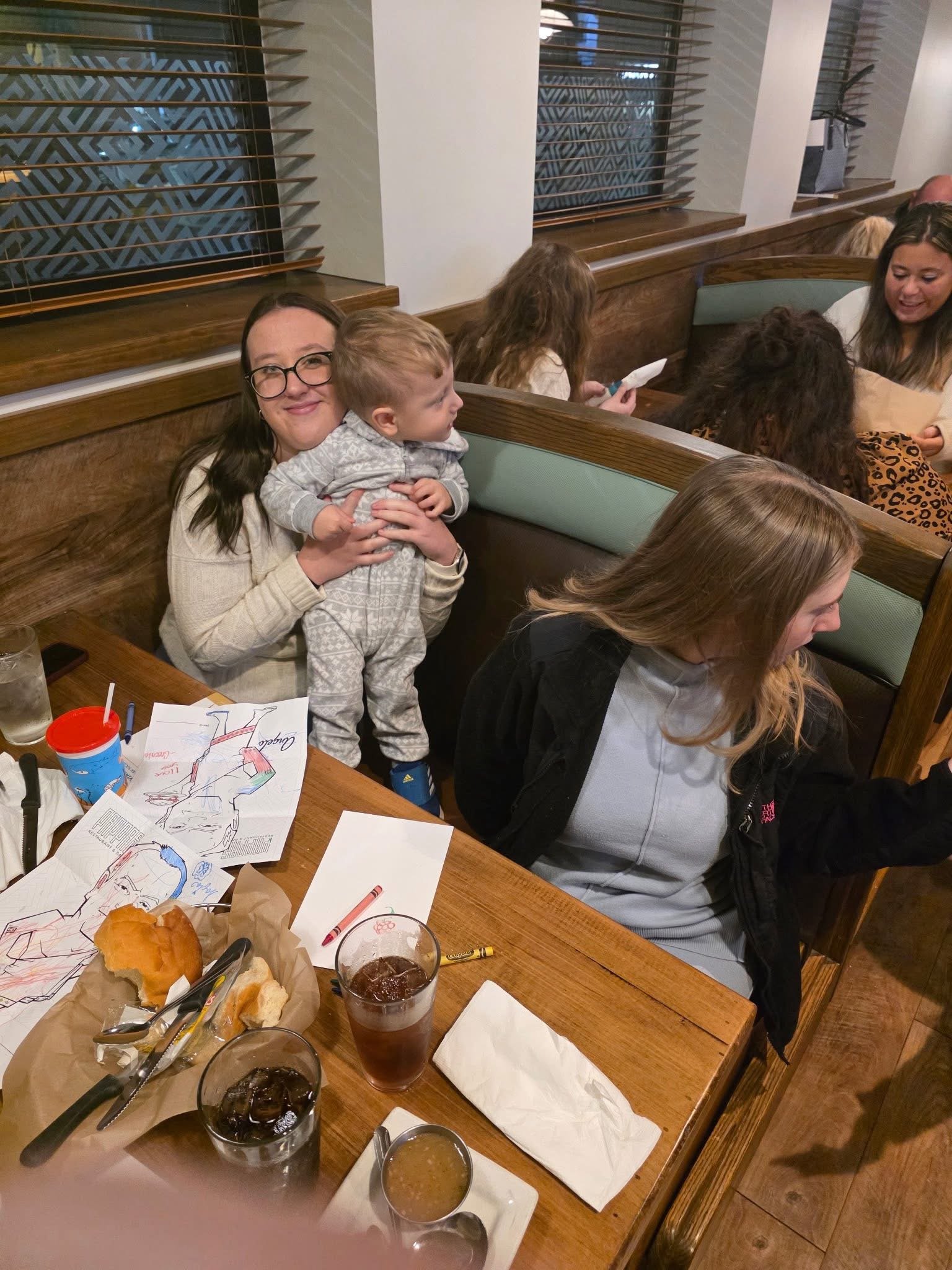 A woman holding a young boy in a restaurant booth, surrounded by other diners, with drinks, scraps of paper, and crayons on the table.