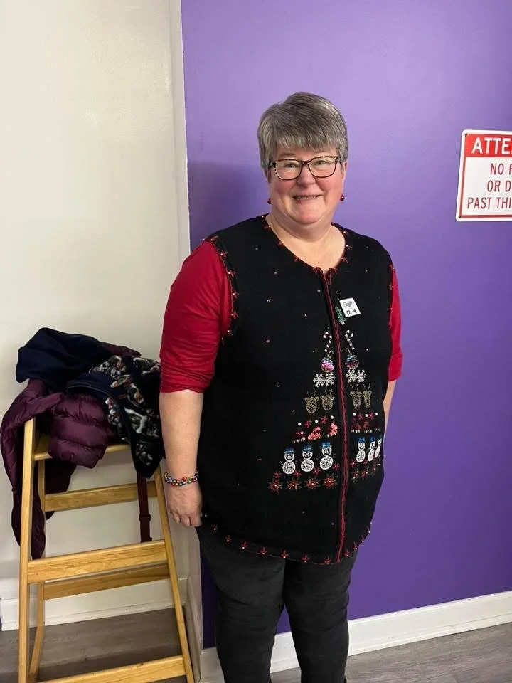 A smiling woman with short gray hair and glasses standing in front of a purple wall. She is wearing a Christmas sweater vest over a red shirt, along with black pants and a colorful beaded bracelet. To her left, a wooden chair holds a purple coat and 