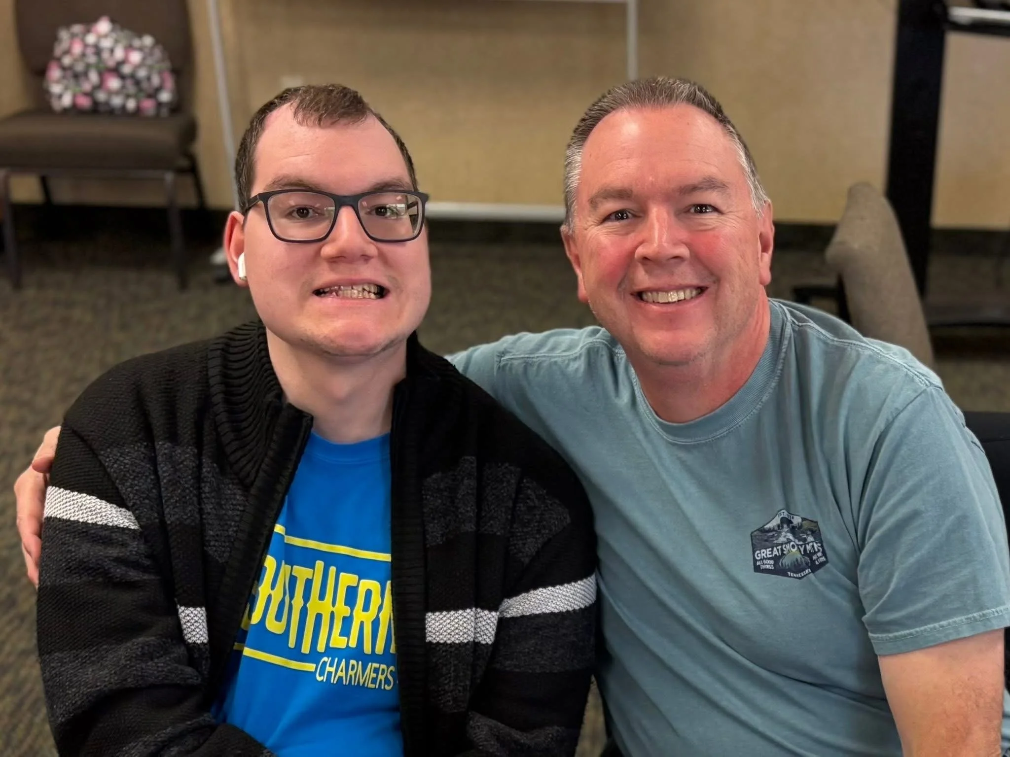 Two men smiling and sitting close together indoors, one wearing glasses and a black jacket, the other in a gray T-shirt.