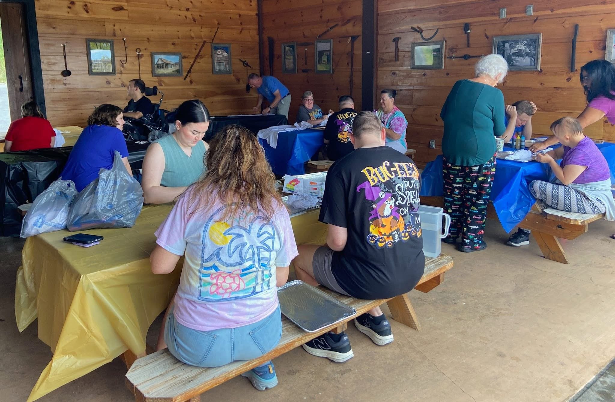 People gathered inside a wooden building, sitting at tables covered with colorful tablecloths, engaging in various activities such as talking, preparing food, or looking down at items on the table.