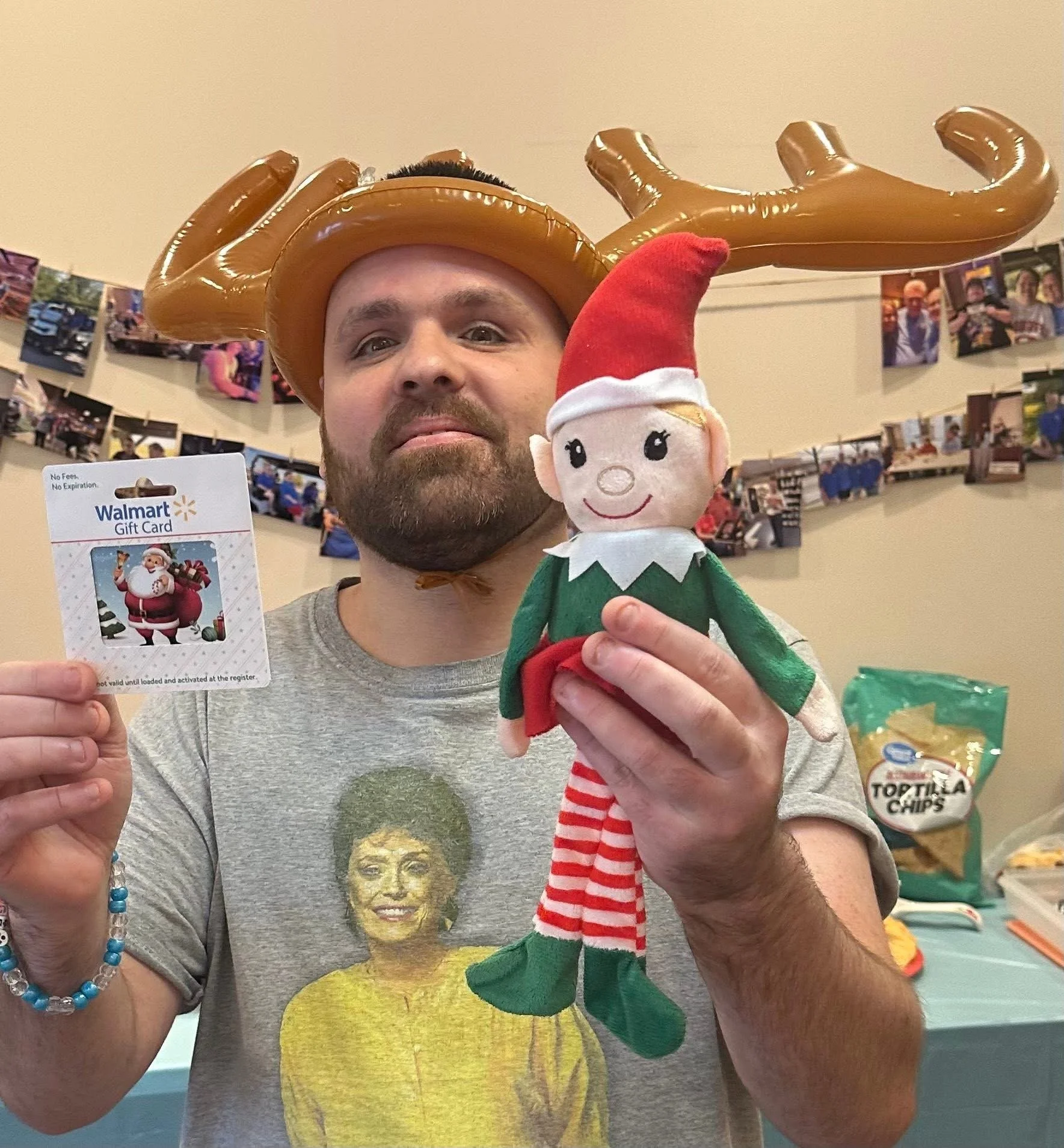 A man wearing a reindeer-shaped balloon hat and holding an elf plush toy. The man is also holding a Walmart gift card featuring Santa Claus. In the background, there are photographs hanging on the wall and a bag of tortilla chips on a table.