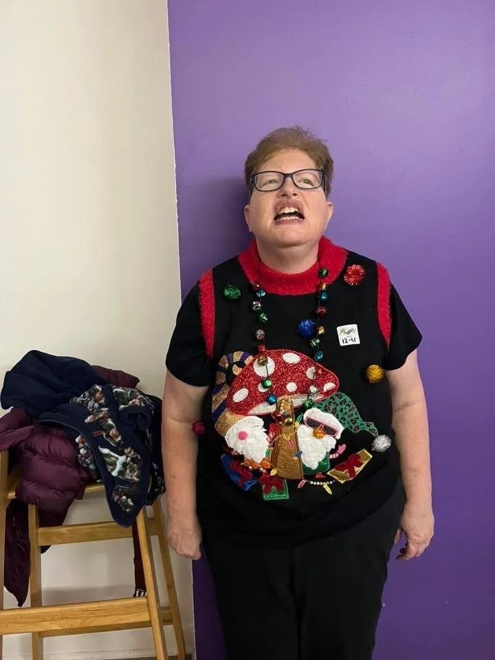 An older woman with glasses and short brown hair making a funny face, standing in front of a purple wall. She is wearing a black Christmas sweater with festive decorations and a necklace with colorful beads. There is a chair with clothes on it to her