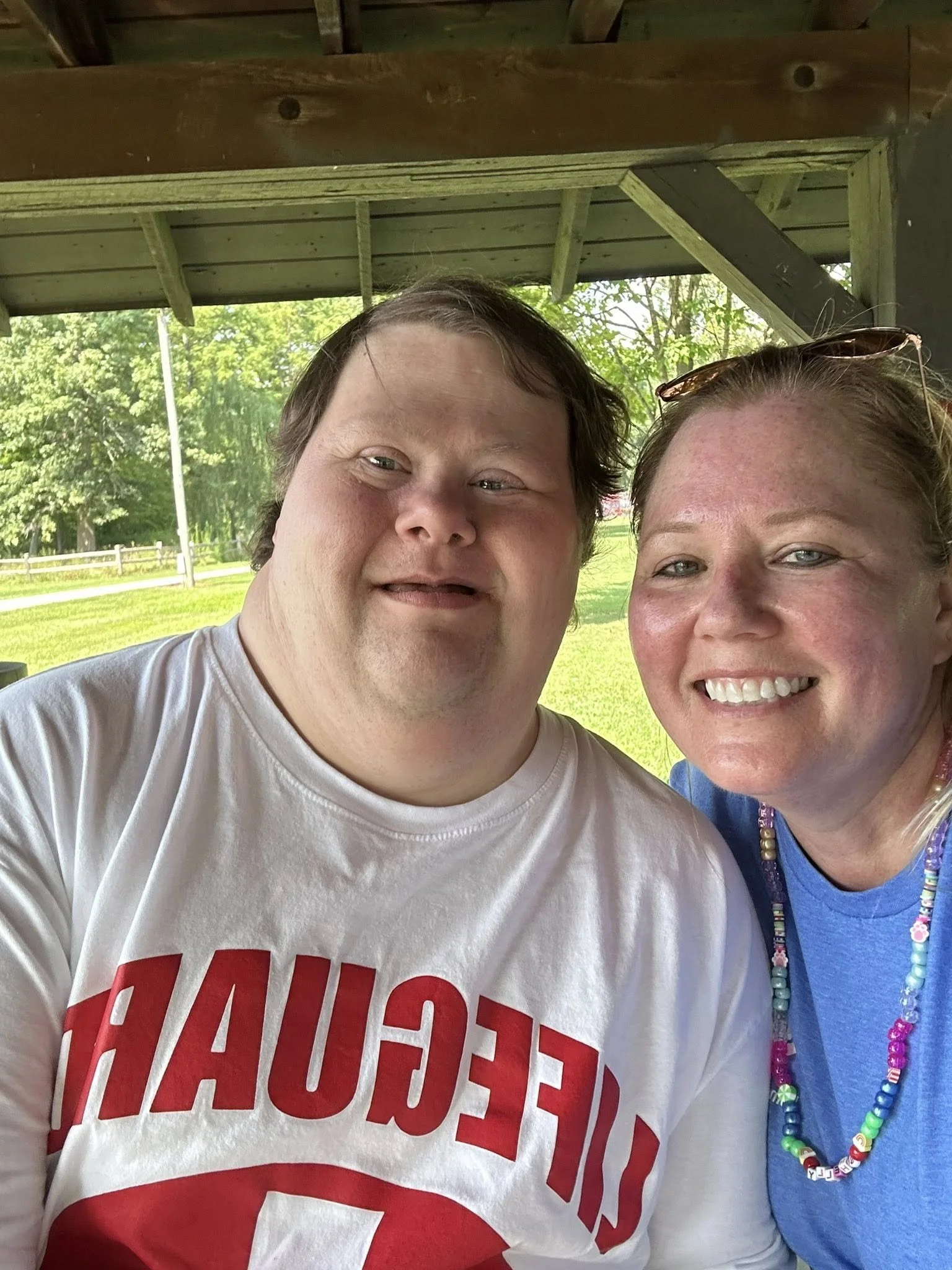 Two smiling people taking a selfie, outdoors under a wooden shelter with green trees and grass in the background.