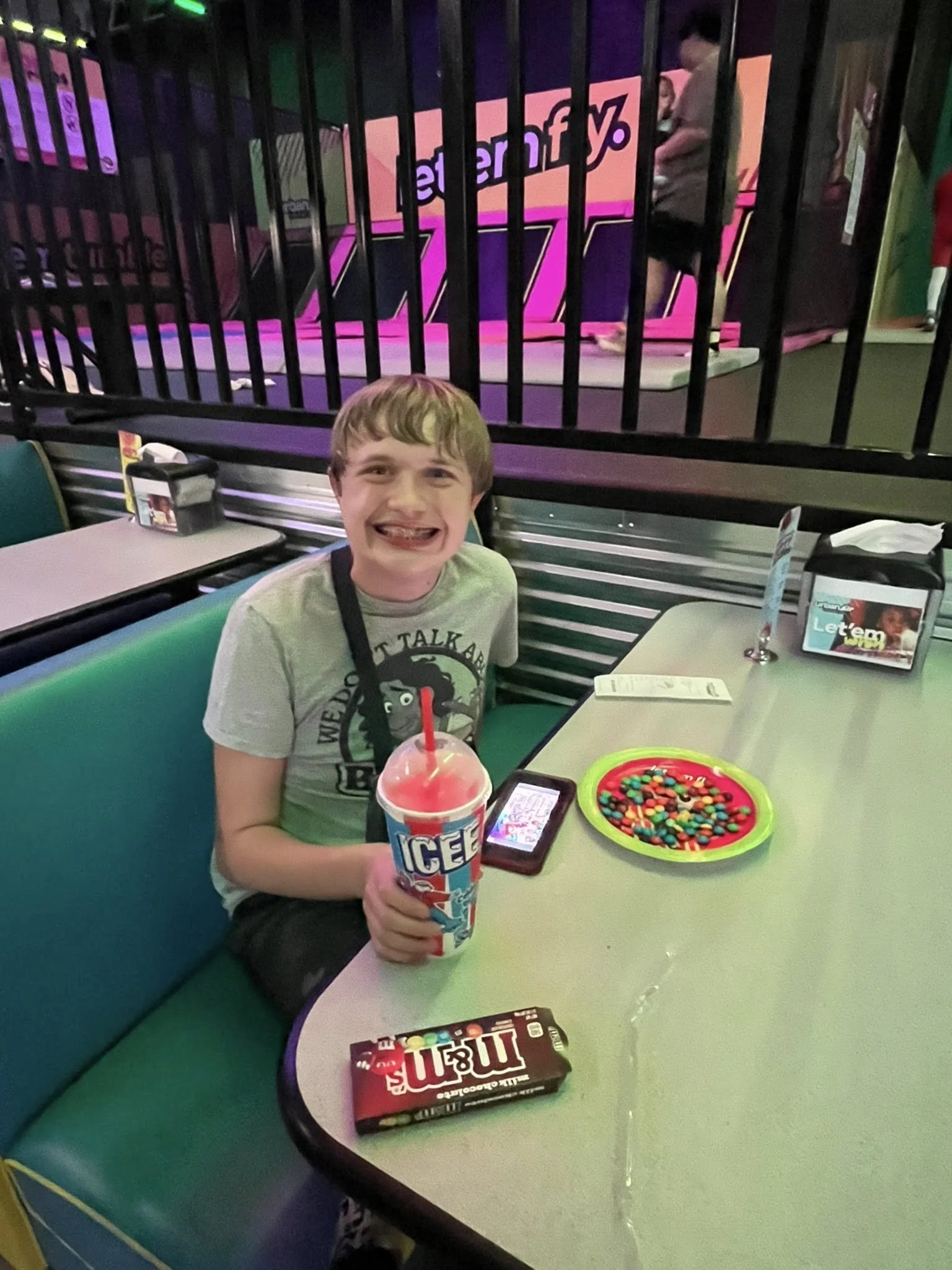 A boy sitting at a table in an arcade, holding a large icee drink, with a mobile phone, a box of M&M's, and a colorful plate of candy or M&Ms on the table. He is smiling broadly and appears to be enjoying his time.