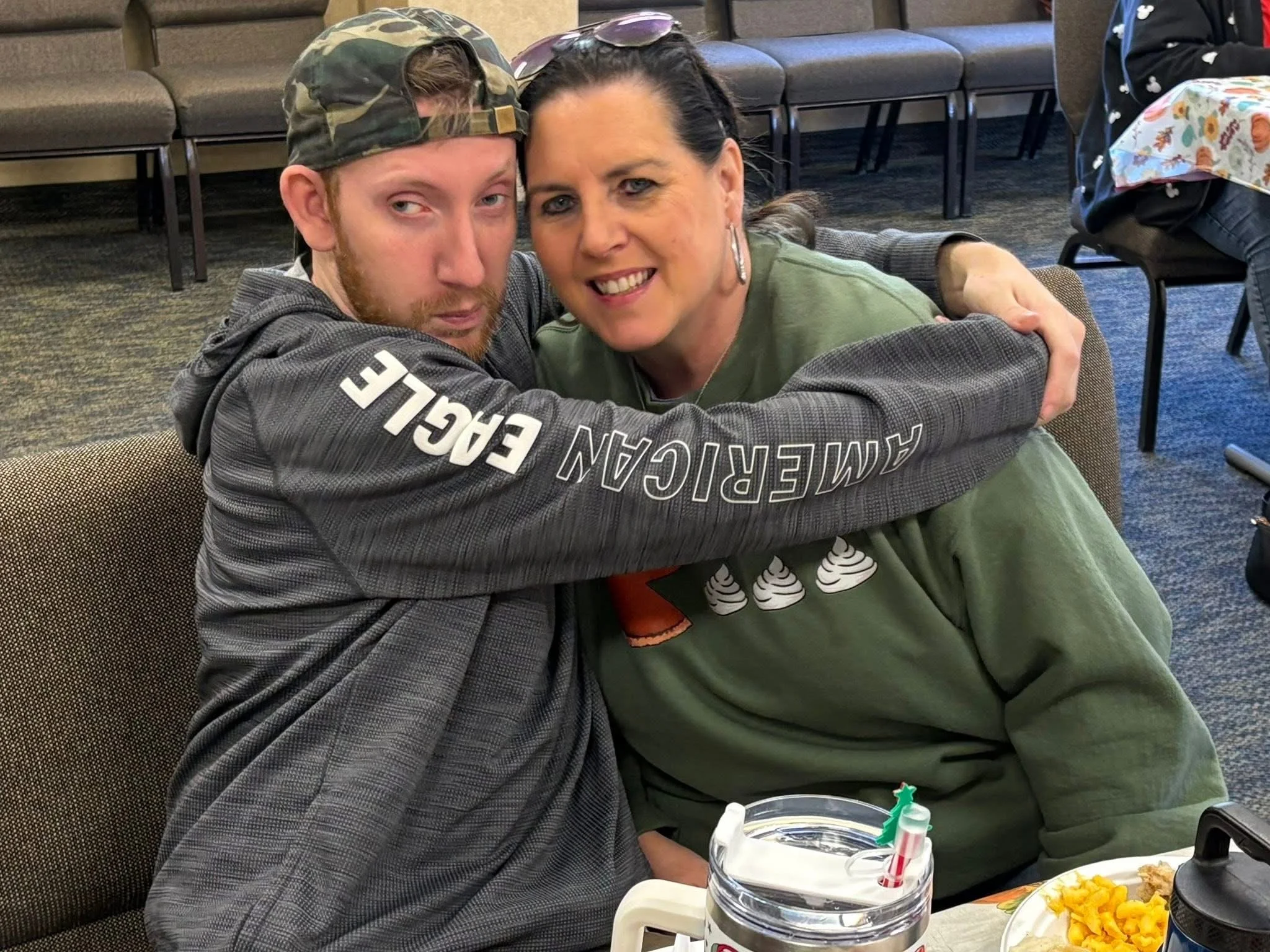 A young man and a woman are hugging and smiling at the camera in a room with chairs and other people. The young man has a beard, a camo hat worn backwards, and is wearing a gray hoodie with 'AMERICAN EAGLE' written on the sleeve. The woman has dark h