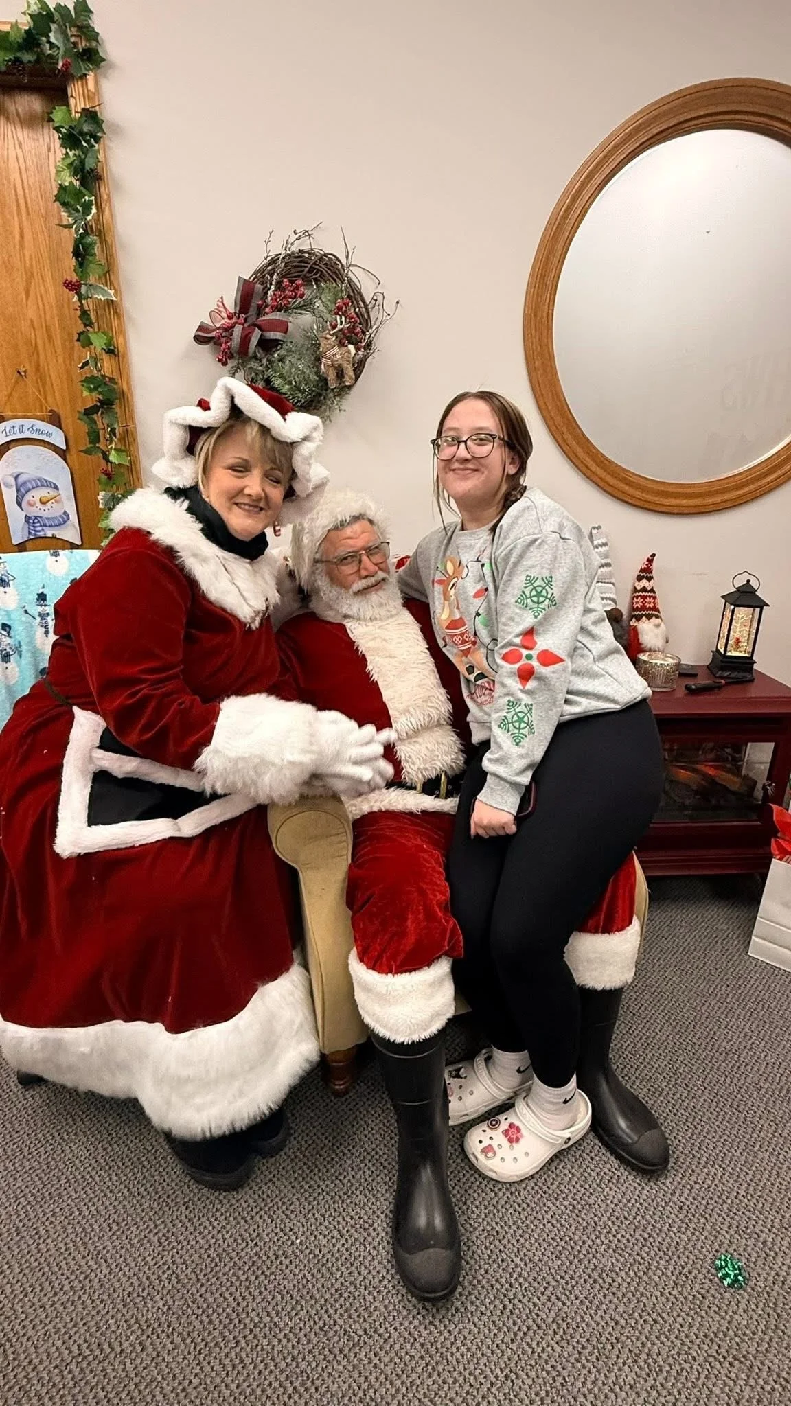 A young girl with glasses posing with Santa Claus and Mrs. Claus indoors during Christmas, with holiday decorations on the wall behind them.