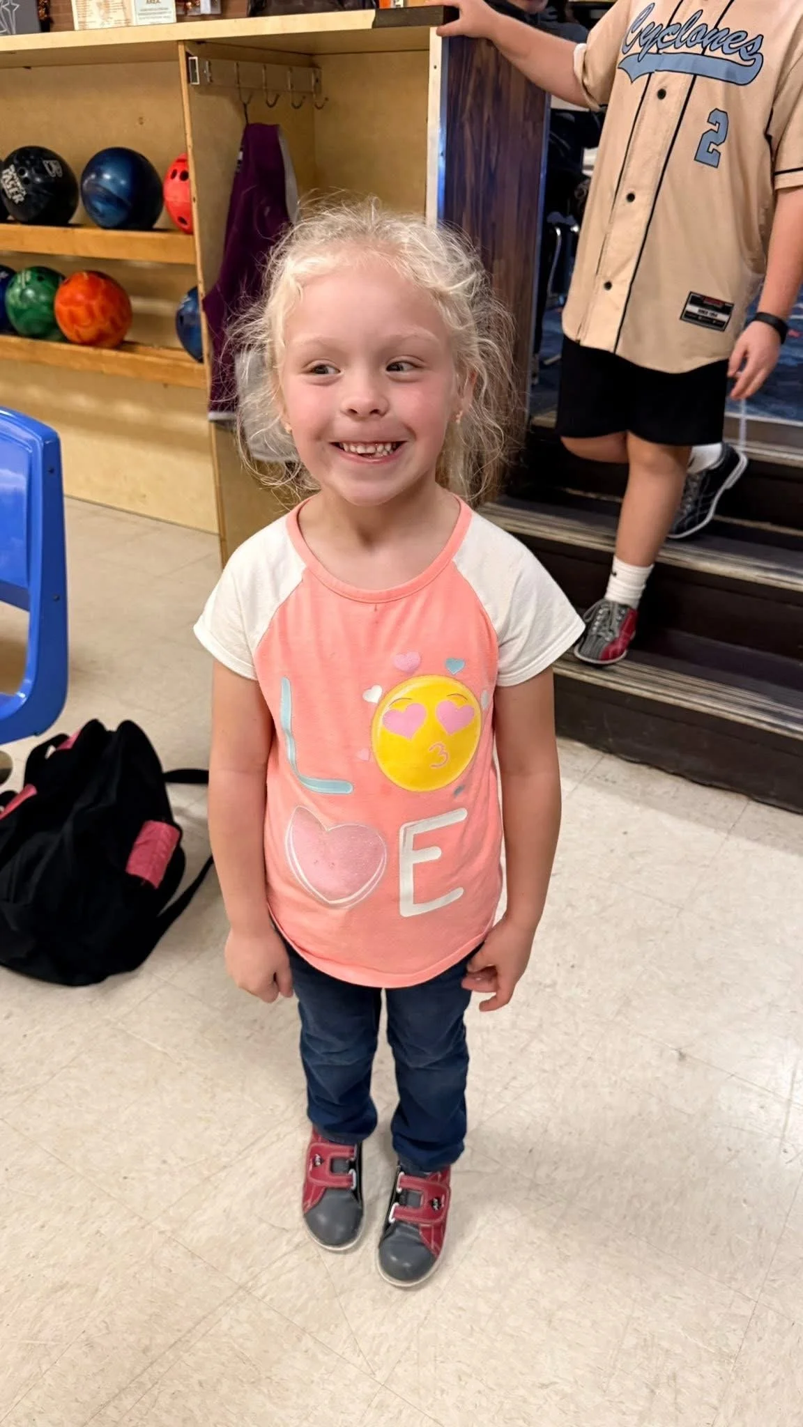 A young girl with curly blonde hair smiling inside a bowling alley, wearing a colorful T-shirt with emoji and letter designs, navy pants, and red and gray sneakers, standing next to a black backpack.