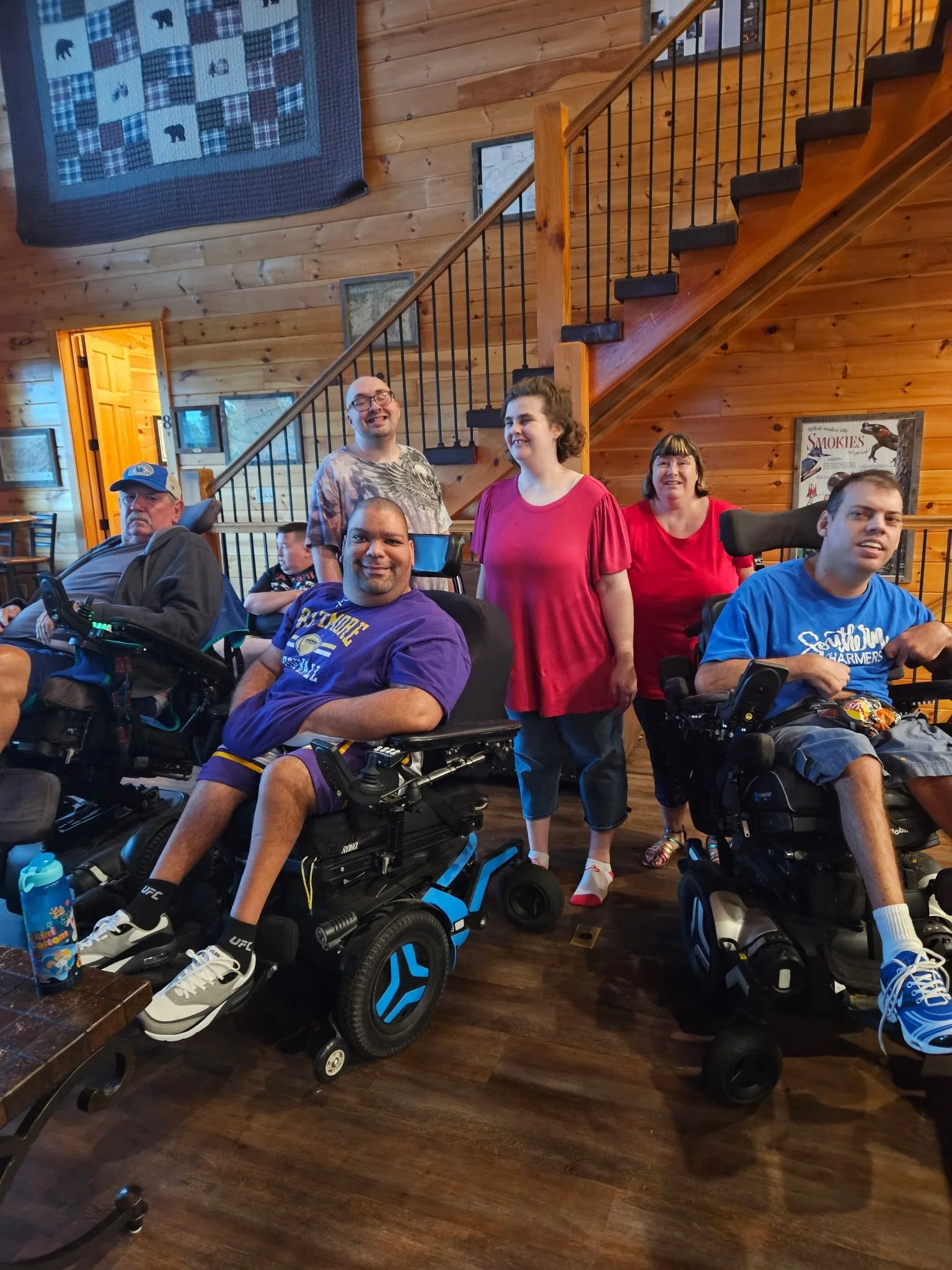 Group of six people, including three in wheelchairs, smiling indoors in a wooden room with a staircase and framed pictures on the wall.