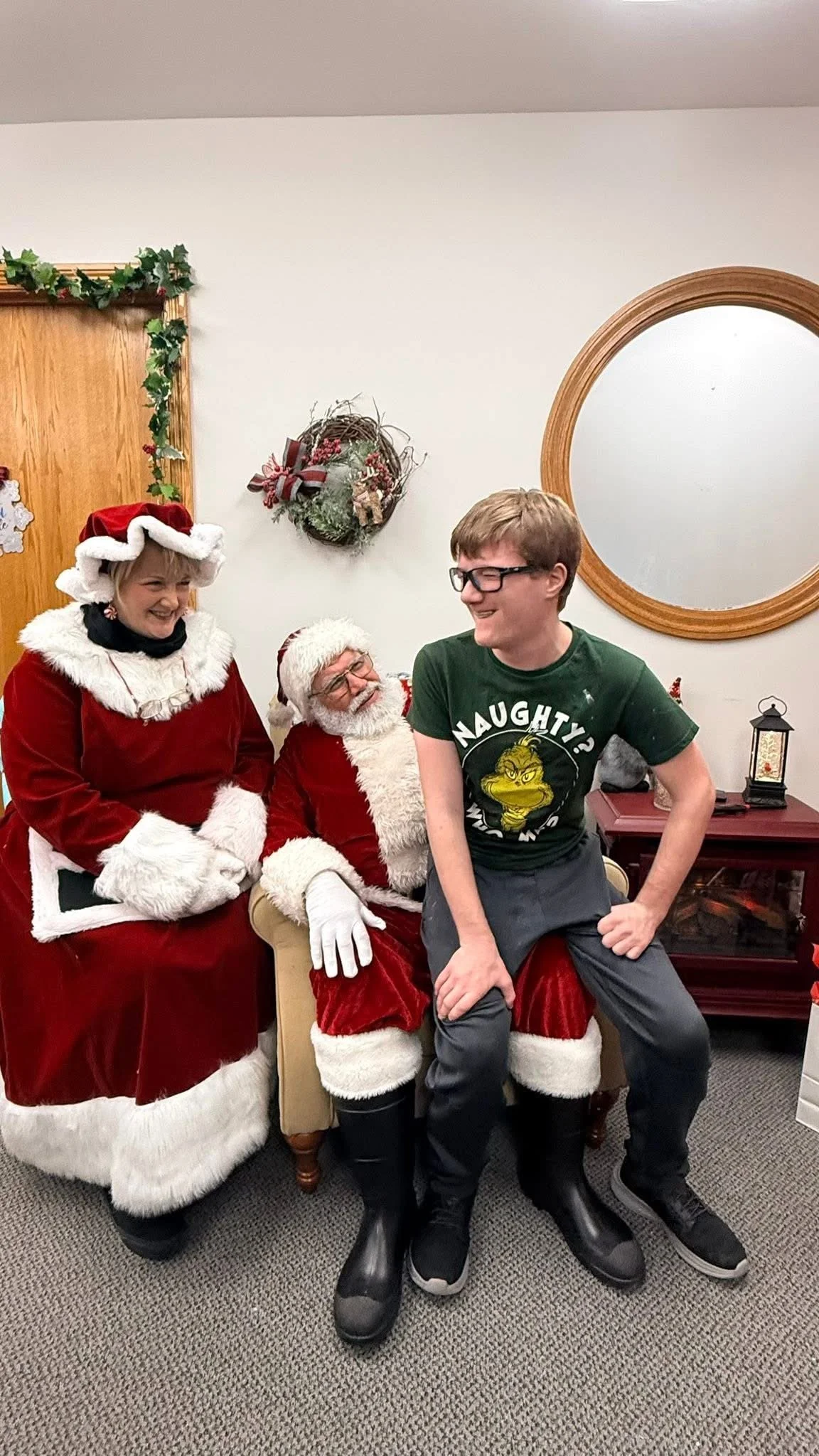 A young man sitting on Santa's lap with an older woman and Santa Claus standing nearby, all dressed in Christmas attire and smiling.