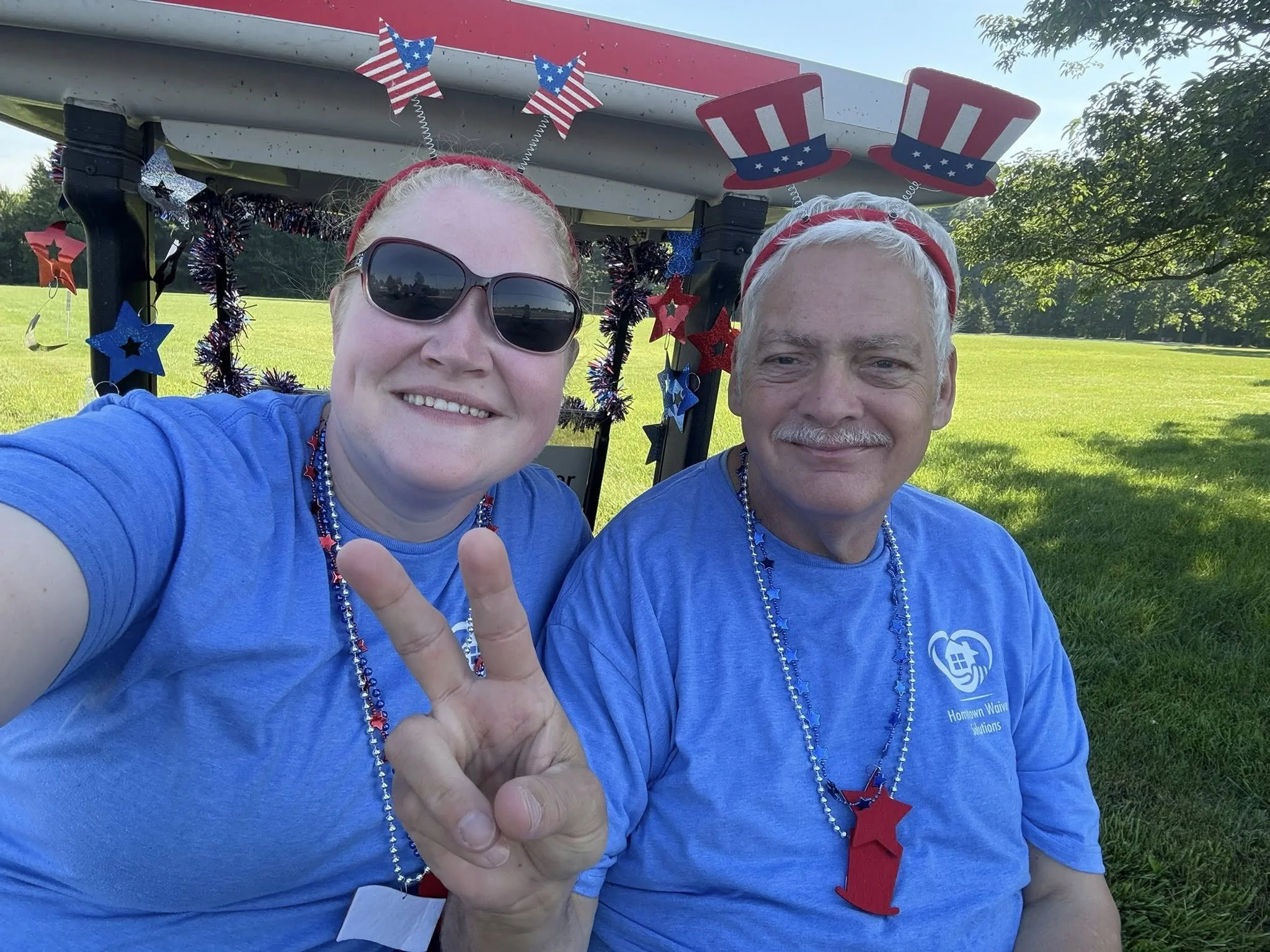 A smiling woman and man wearing blue T-shirts and patriotic accessories, sitting on grass. The woman is making a peace sign. They are under a decorated float with American-themed stars and hats.