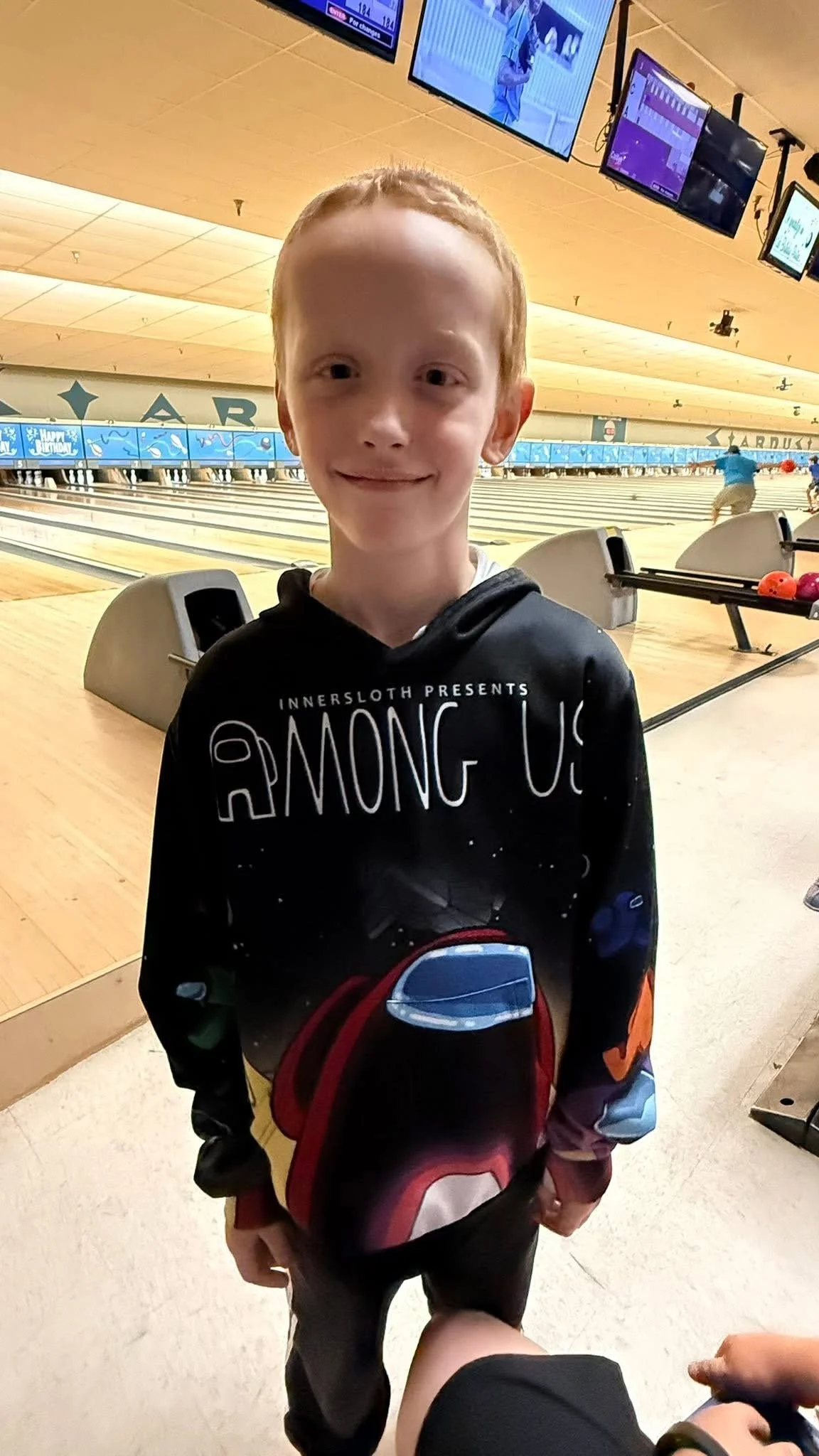 A young boy with red hair smiling at the camera in a bowling alley, wearing a black hoodie with "Among Us" themed design.
