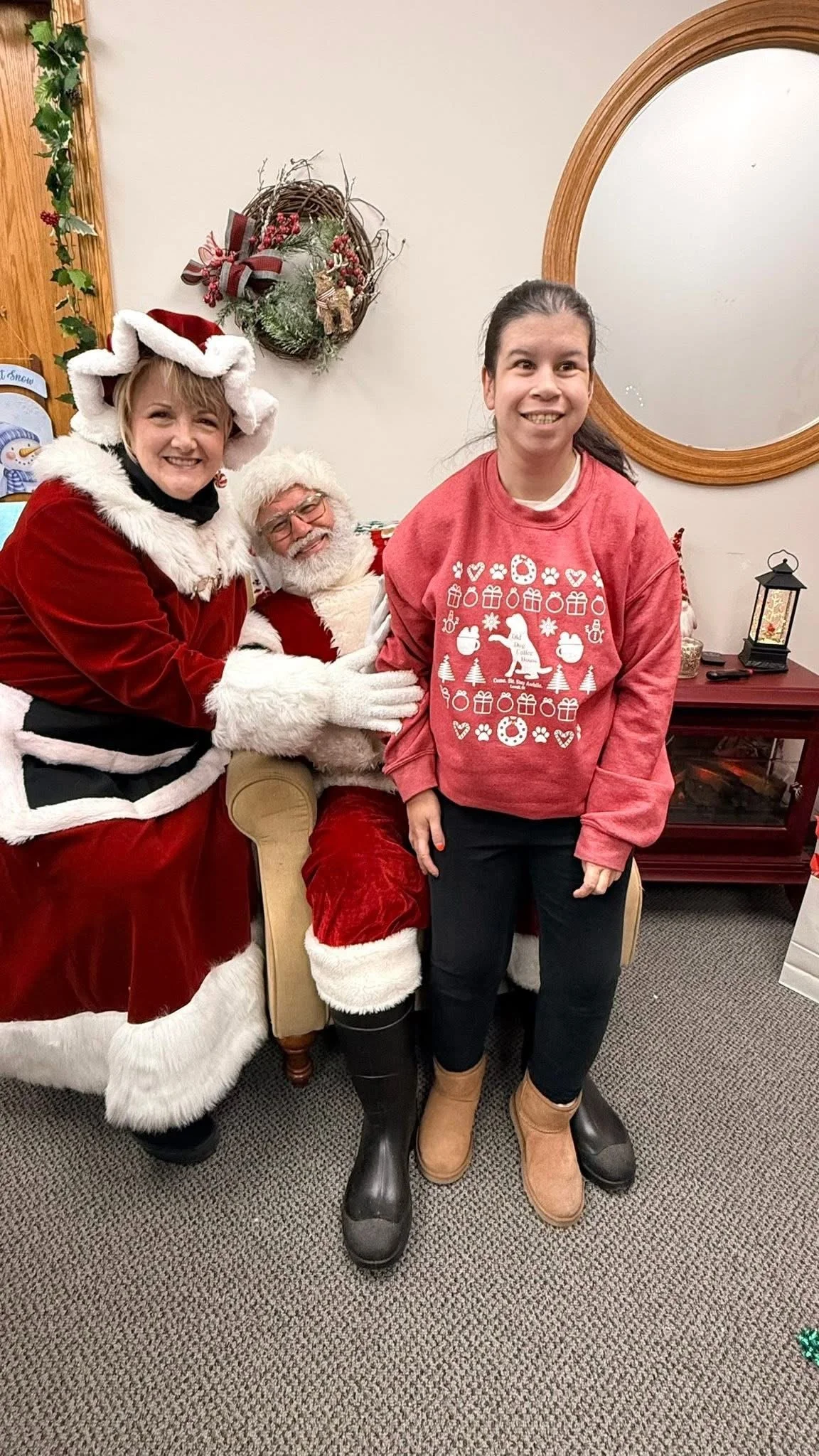 A woman dressed as Mrs. Claus, a man dressed as Santa Claus, and a girl are smiling and posing for a photo indoors decorated for Christmas. Mrs. Claus is sitting on the armrest of Santa's chair, and the girl stands in front of Santa, all smiling.