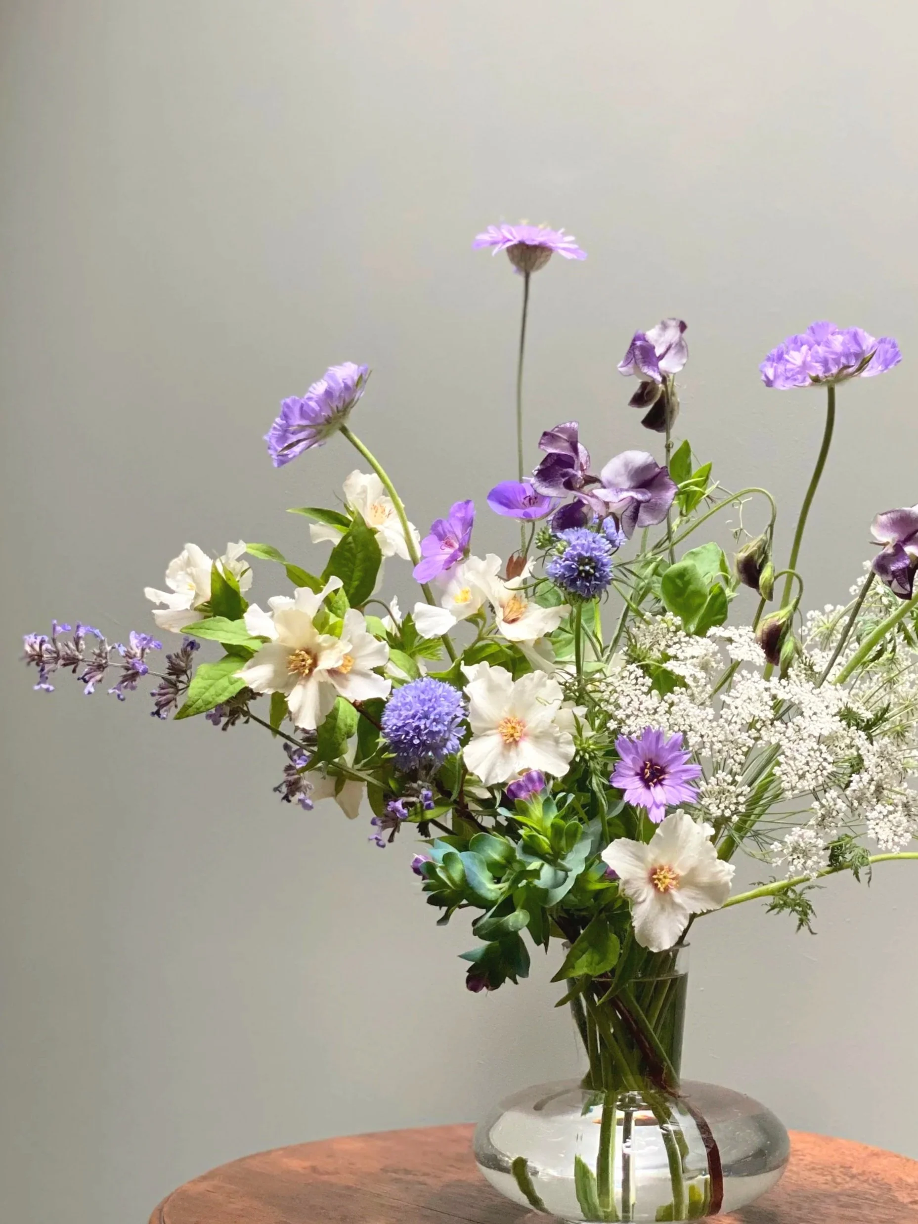A glass vase with a bouquet of natural, seasonal, mixed purple, white, and blue flowers on a wooden table against a plain background.