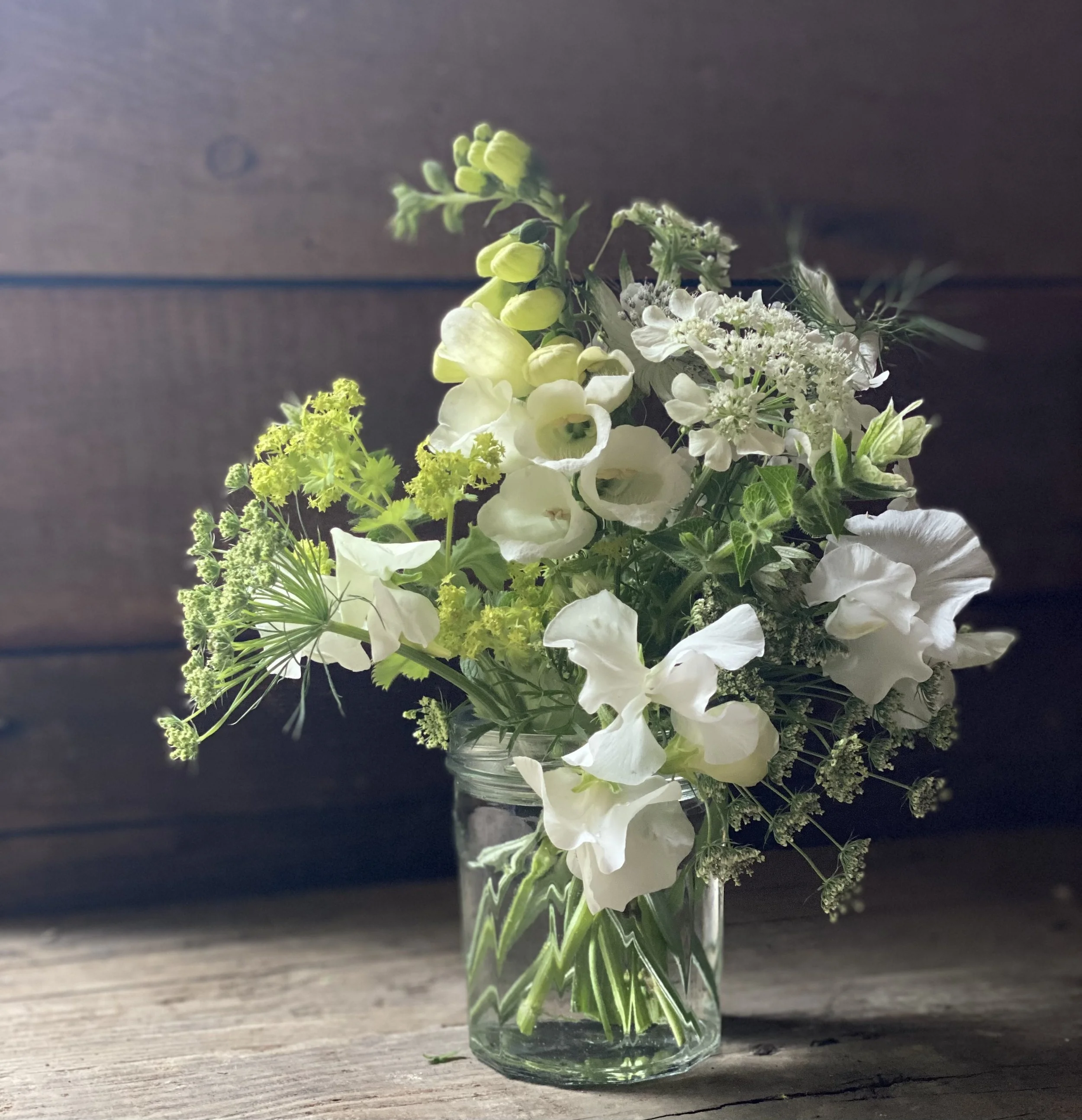 A jam jar posy of white and green flowers, including foxgloves on a wooden surface, with a dark wooden background.