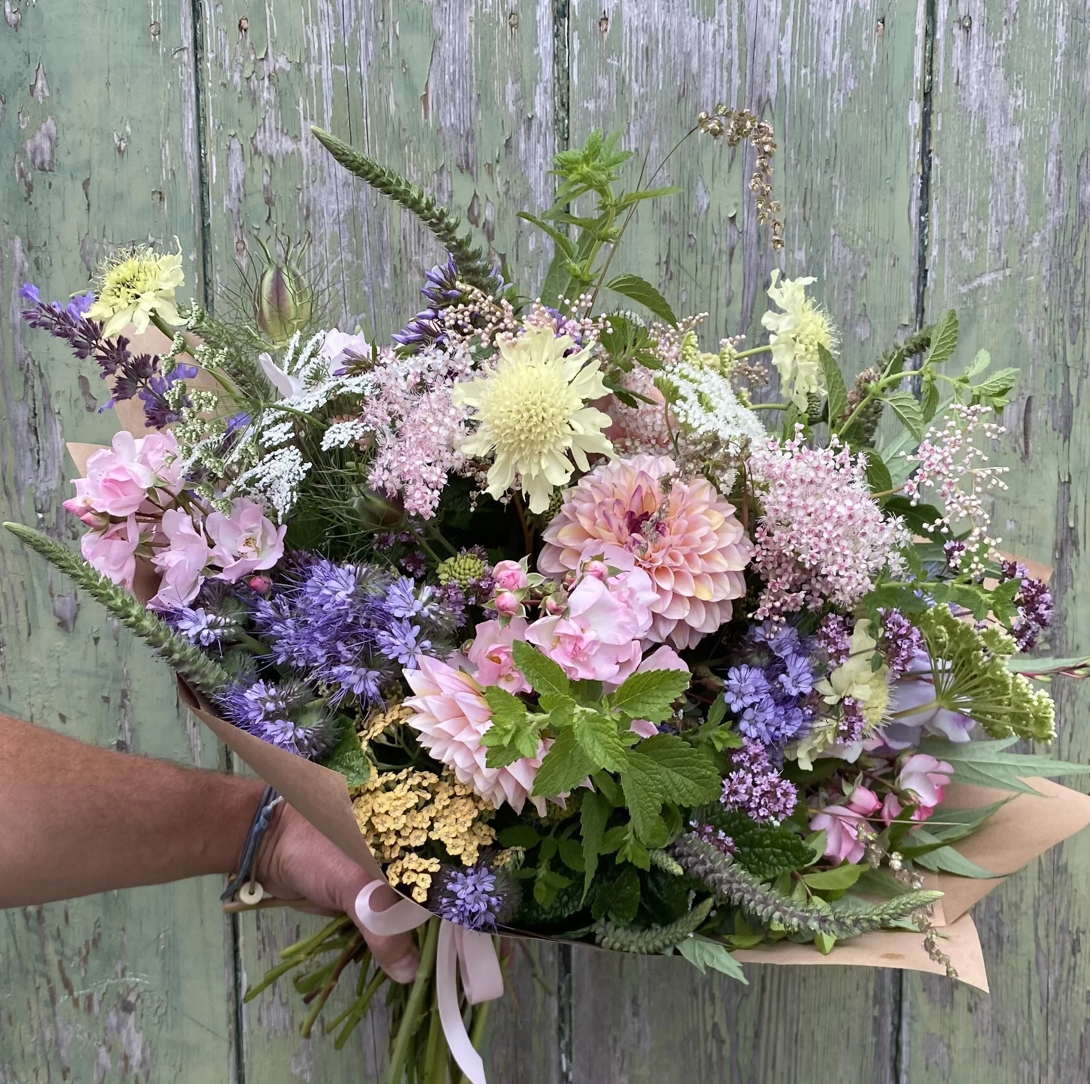 Hand holding a large bouquet of mixed pastel seasonal flowers against a weathered green wooden background.