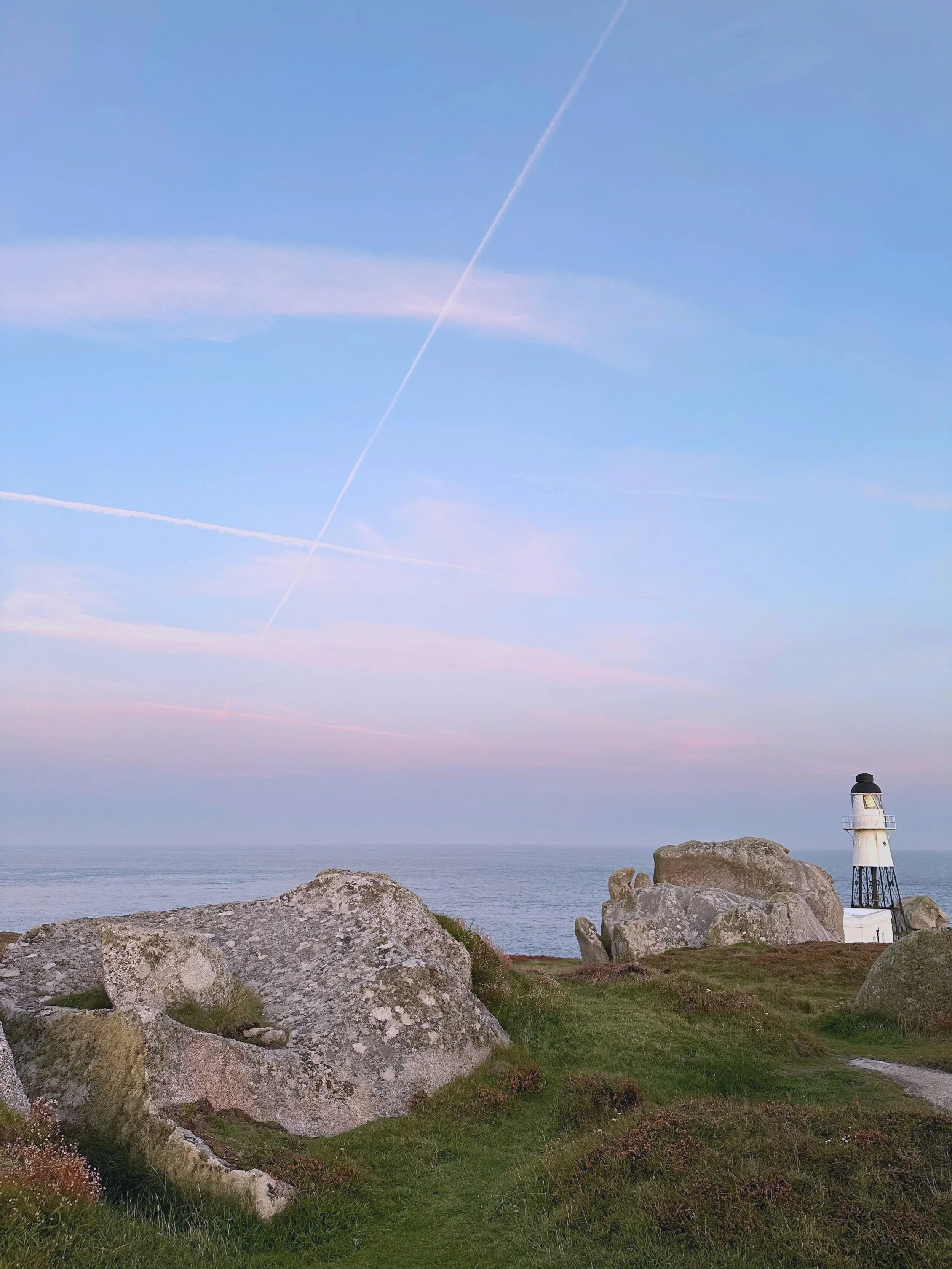 A lighthouse on the Isles of Scilly on a grassy cliff overlooking the ocean with large rocks in the foreground and jet contrails in pink and blue sky.
