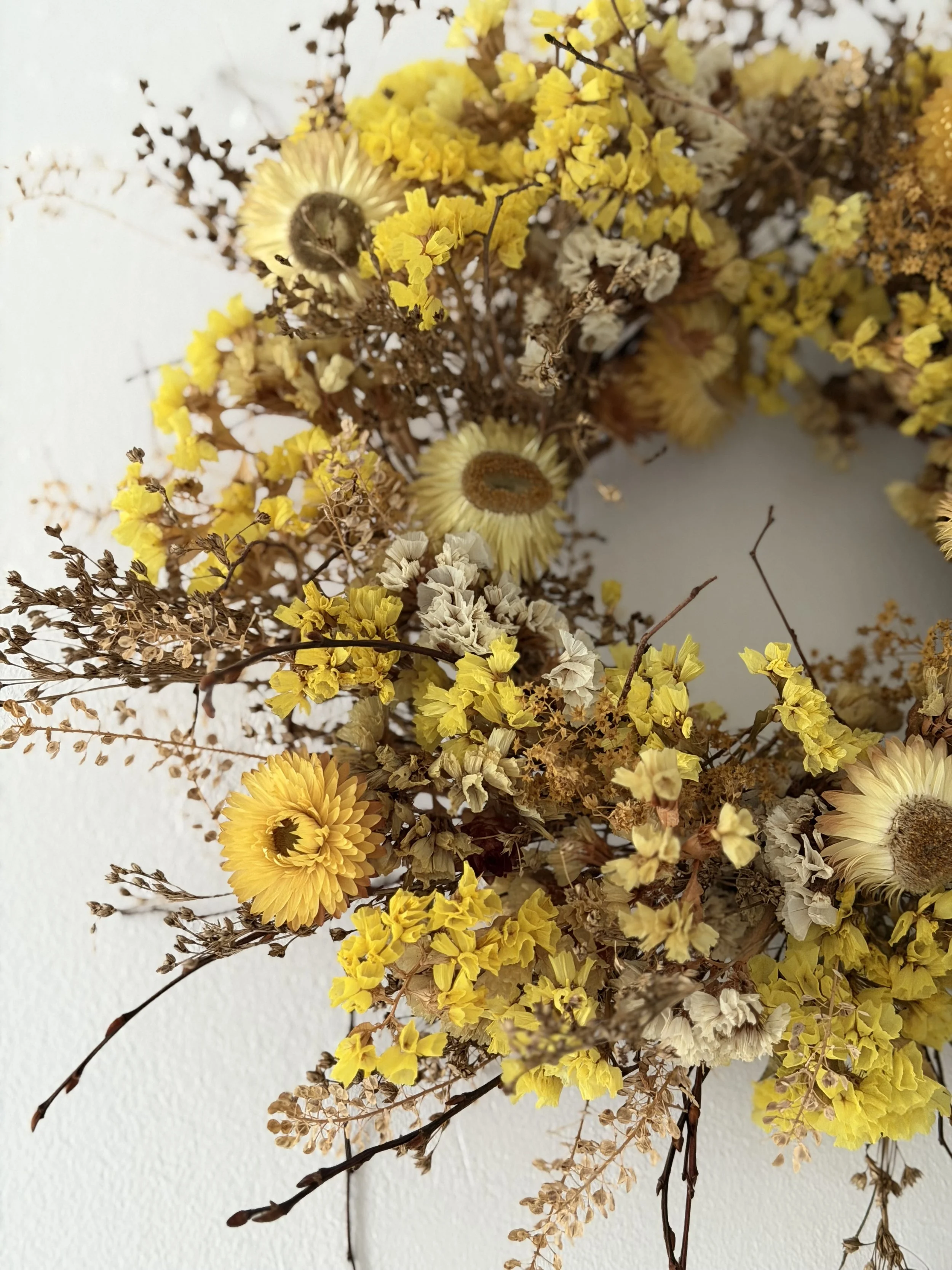 A wreath made of dried yellow, white, and brown flowers and foliage hanging on a white wall.