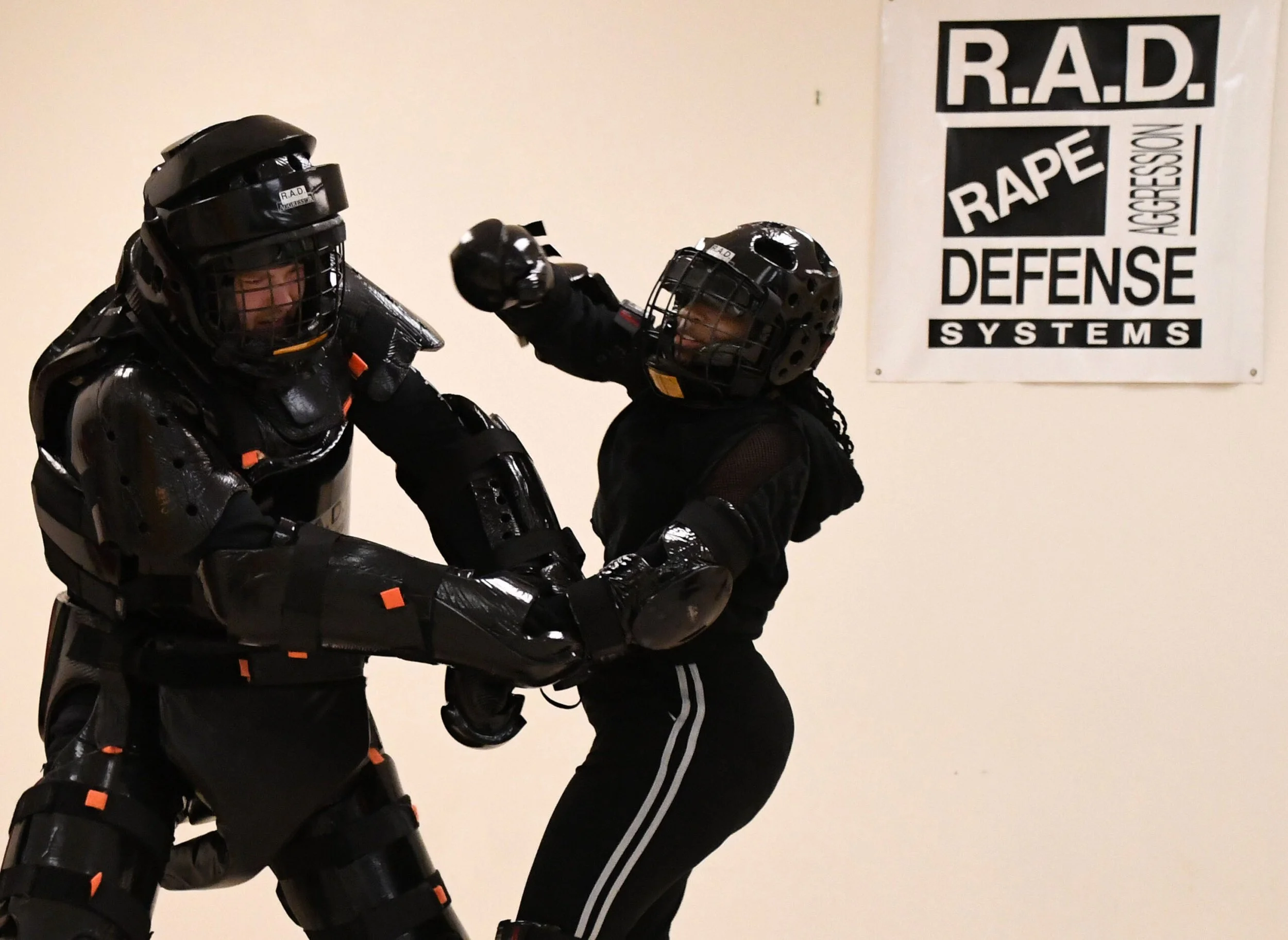 Two individuals wearing combat gear practicing self-defense techniques in a room. One person is in full protective armor, and the other is wearing a helmet and gloves, engaged in a training move near a wall with a sign that says "R.A.D. Rape Defense Systems."