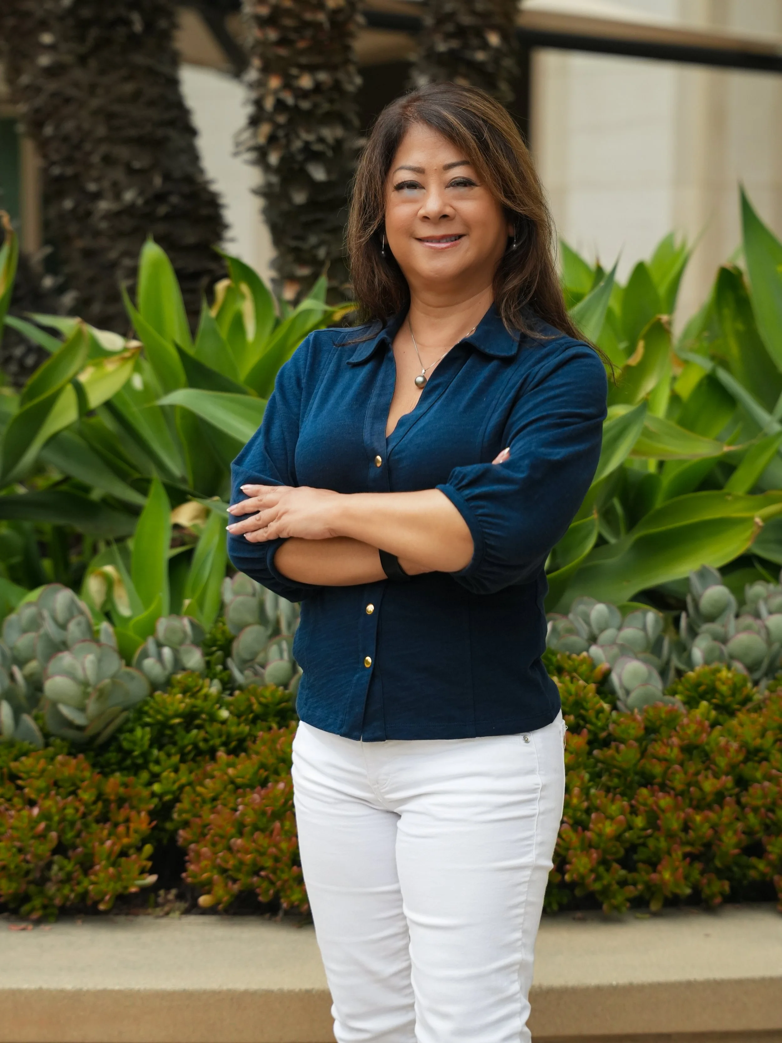 A woman with dark brown hair standing outdoors in front of green plants and bushes, smiling with arms crossed, wearing a navy blue button-up shirt and white pants.