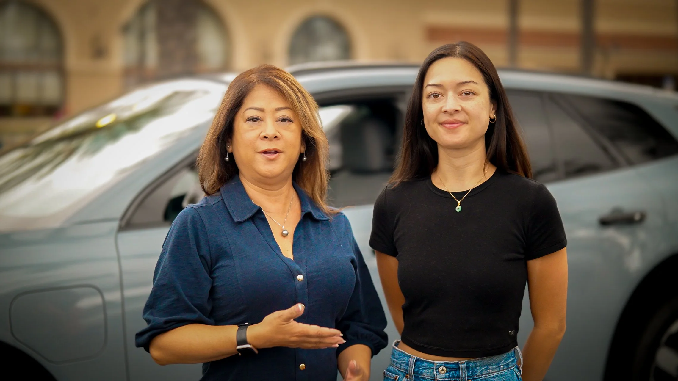 Two women standing in front of a gray car, outdoors with brick buildings in the background. The woman on the left has shoulder-length brown hair and is wearing a blue shirt with pearl jewelry. The woman on the right has long dark hair and is wearing a black t-shirt with a necklace and denim jeans.