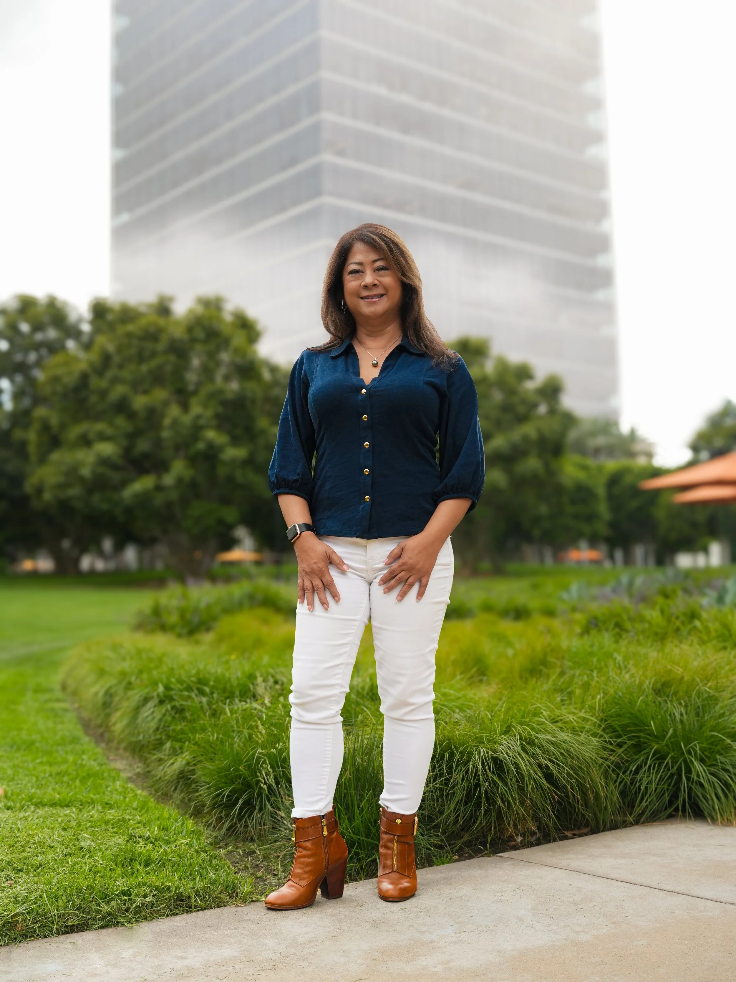 A woman standing on a sidewalk in a park with green grass and trees, wearing a navy blue blouse, white pants, and brown ankle boots, with a modern glass building in the background.
