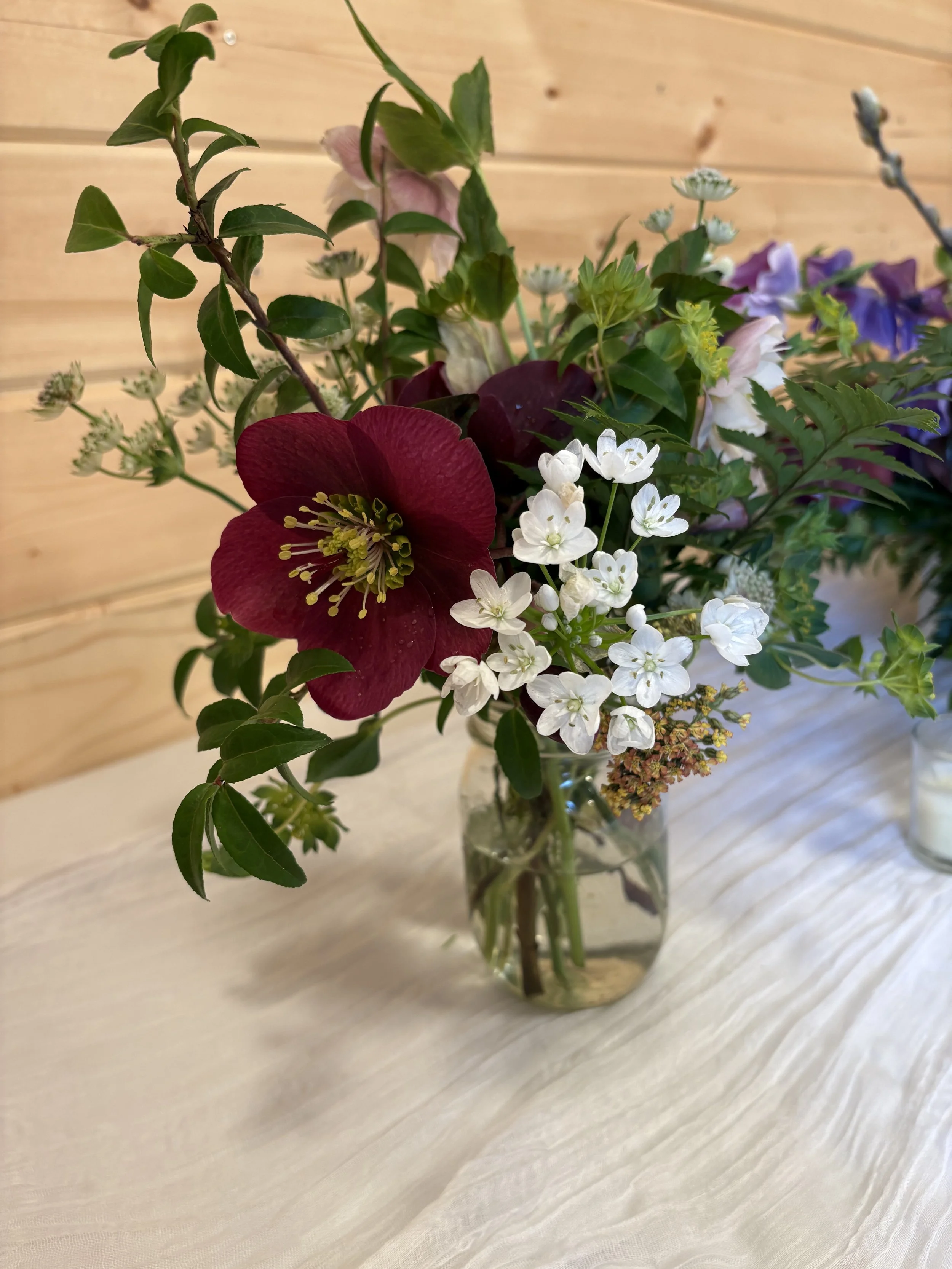 A glass vase filled with a bouquet of colorful flowers, including a large dark red flower, small white blossoms, and various green leaves, placed on a light wooden surface with a wooden wall background.