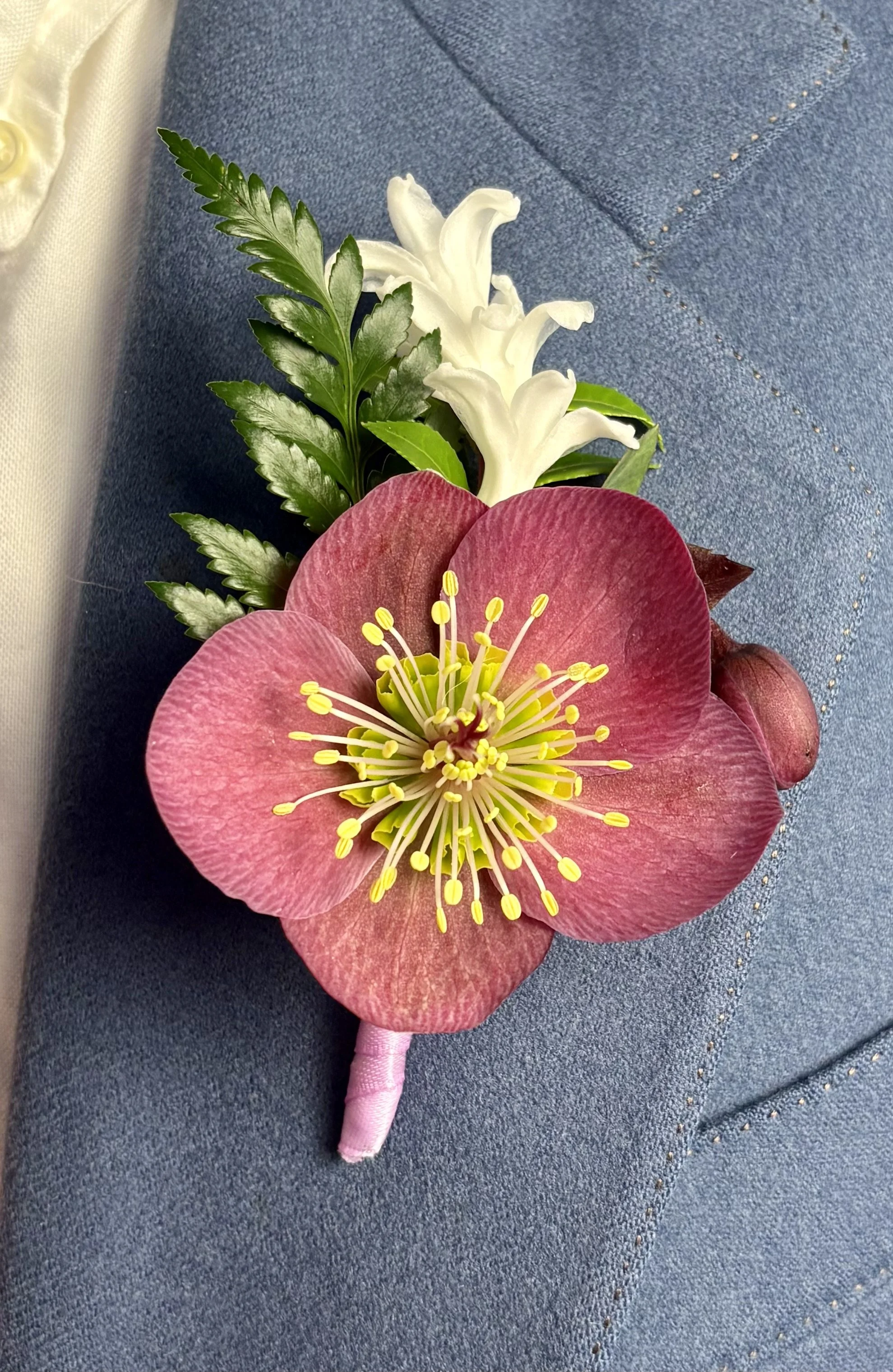 Close-up of a boutonniere with a pink flower, white hyacinth, green leaves, and fern, resting on a blue fabric surface.