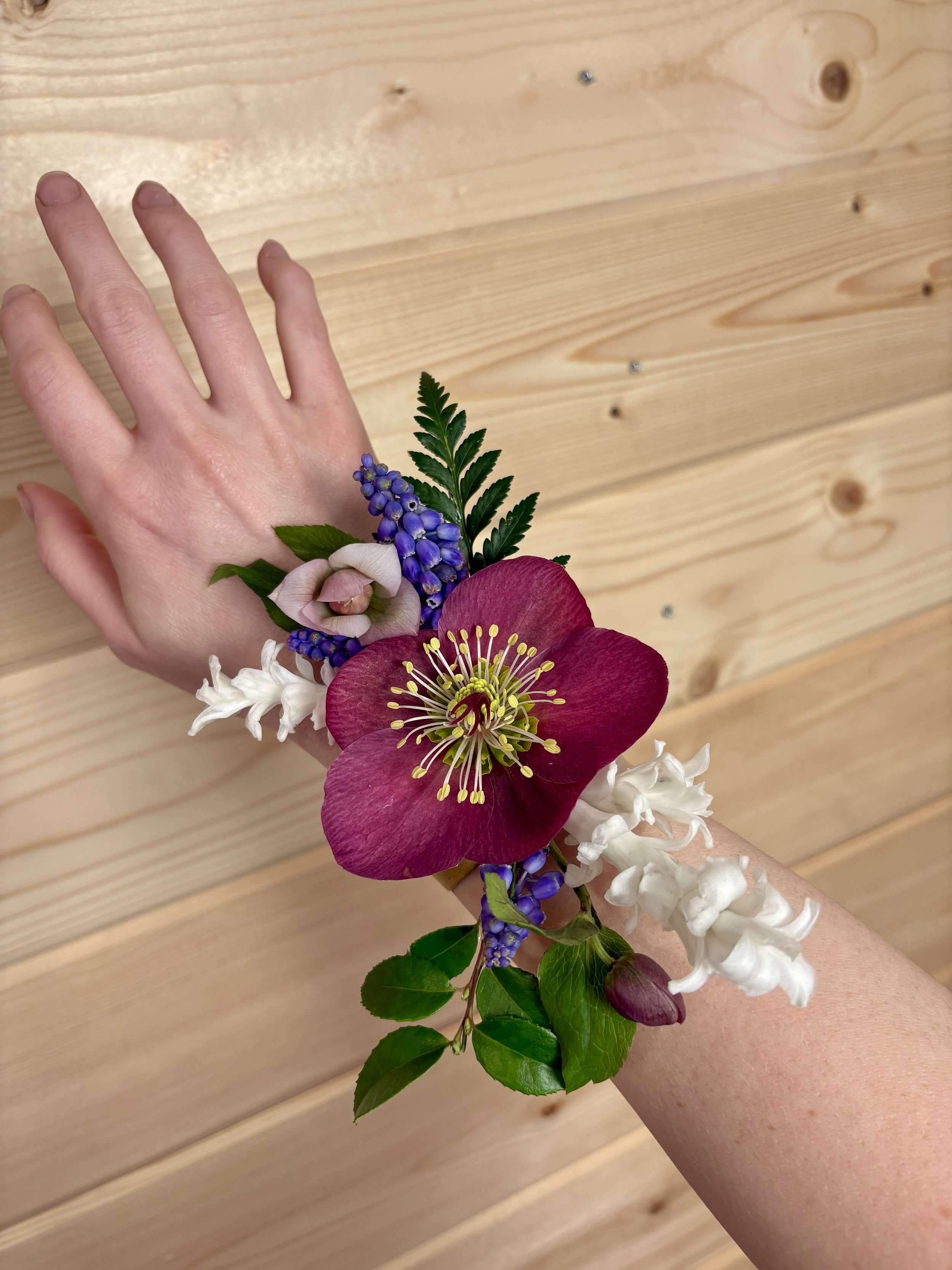 A person's left wrist adorned with a colorful floral corsage, featuring a large pink flower, purple grape hyacinths, white hyacinths, and green leaves, against a background of light wooden planks.
