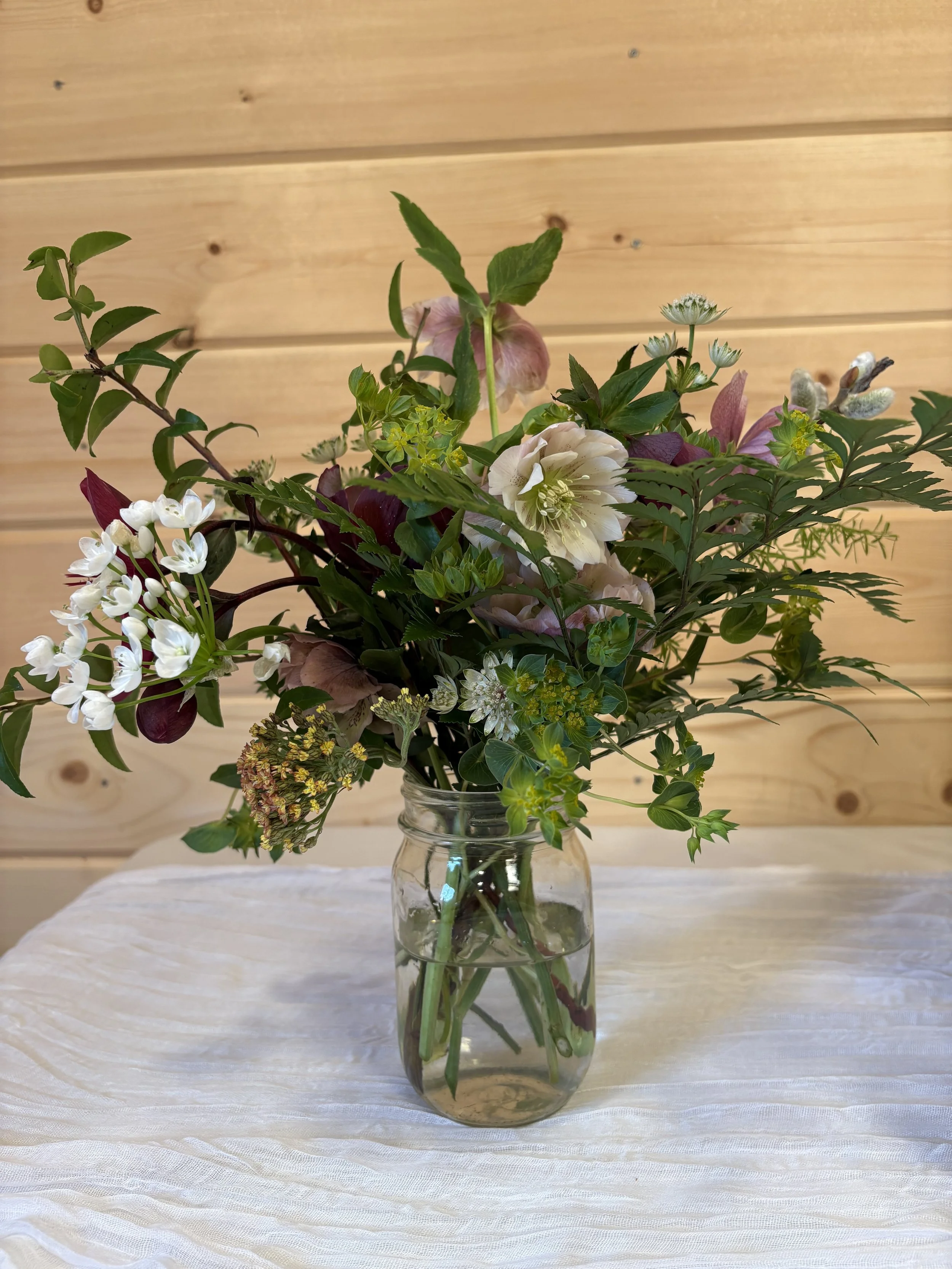 A bouquet of various flowers and greenery in a clear glass vase on a white tablecloth in front of a wooden wall.