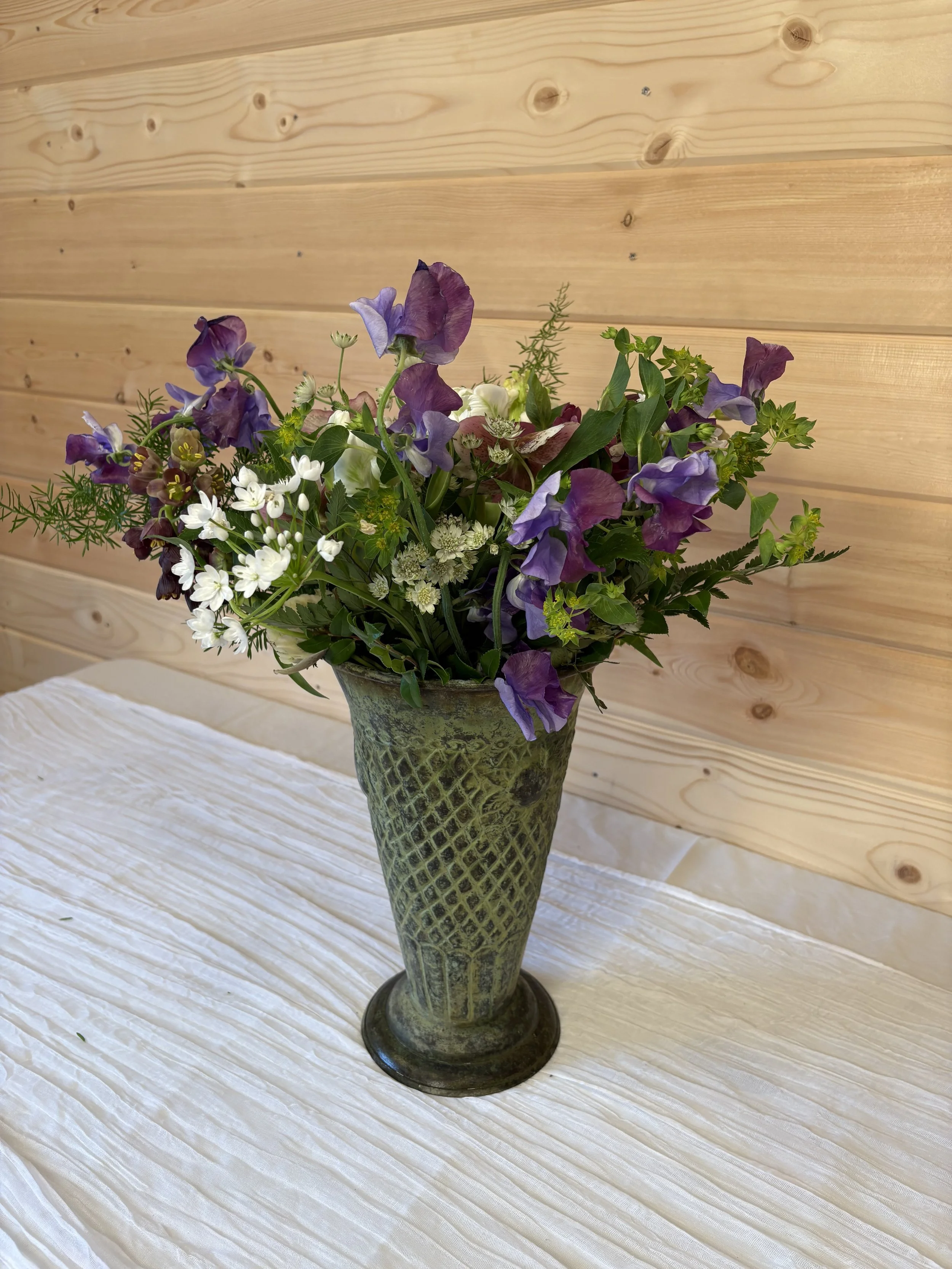 A vase with a bouquet of mixed flowers, including purple, white, and green blooms, placed on a white cloth table with a wooden wall background.