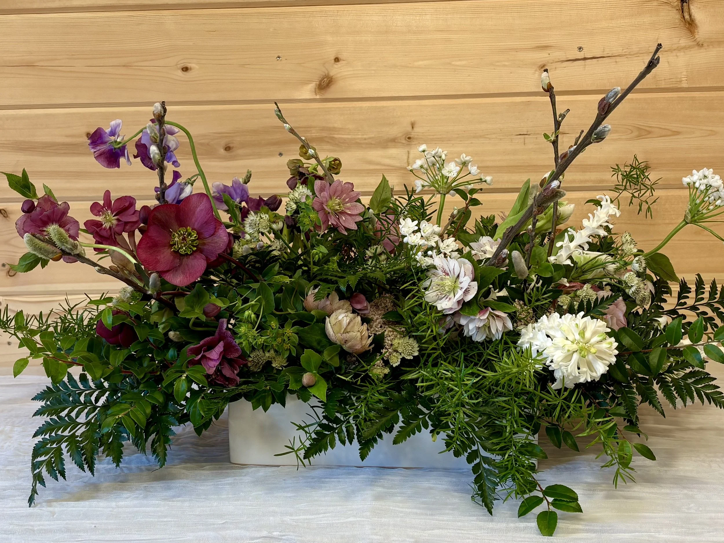 Flower arrangement with various pink, white, and purple flowers, green foliage, and branches in a white rectangular vase on a white table with a wood-paneled wall background.