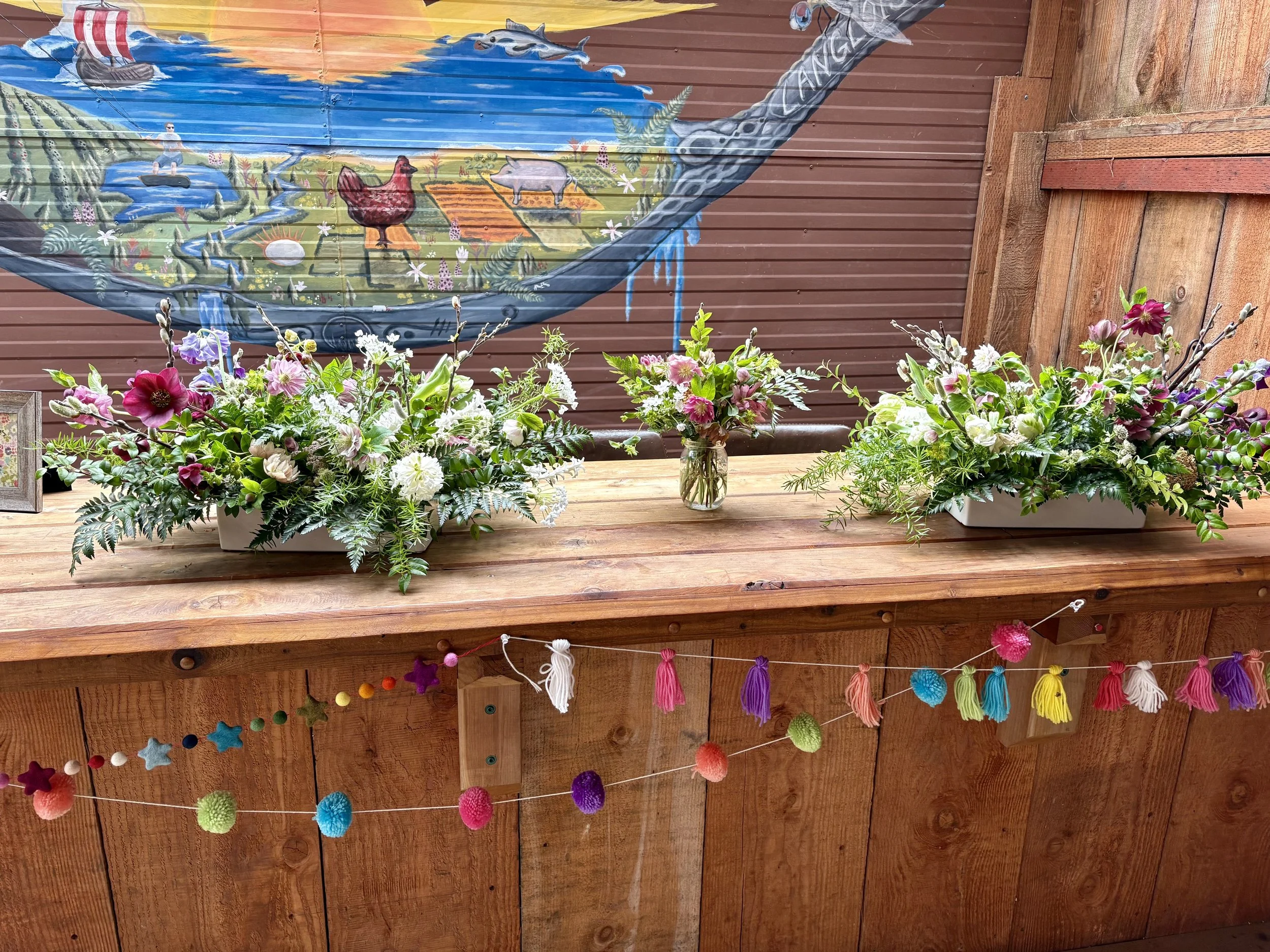 Three floral arrangements with pink, white, purple, and green flowers on a wooden counter, with colorful felt pom-poms and star decorations hanging below, and a colorful painted mural of a boat, water, animals, and a pig in the background.