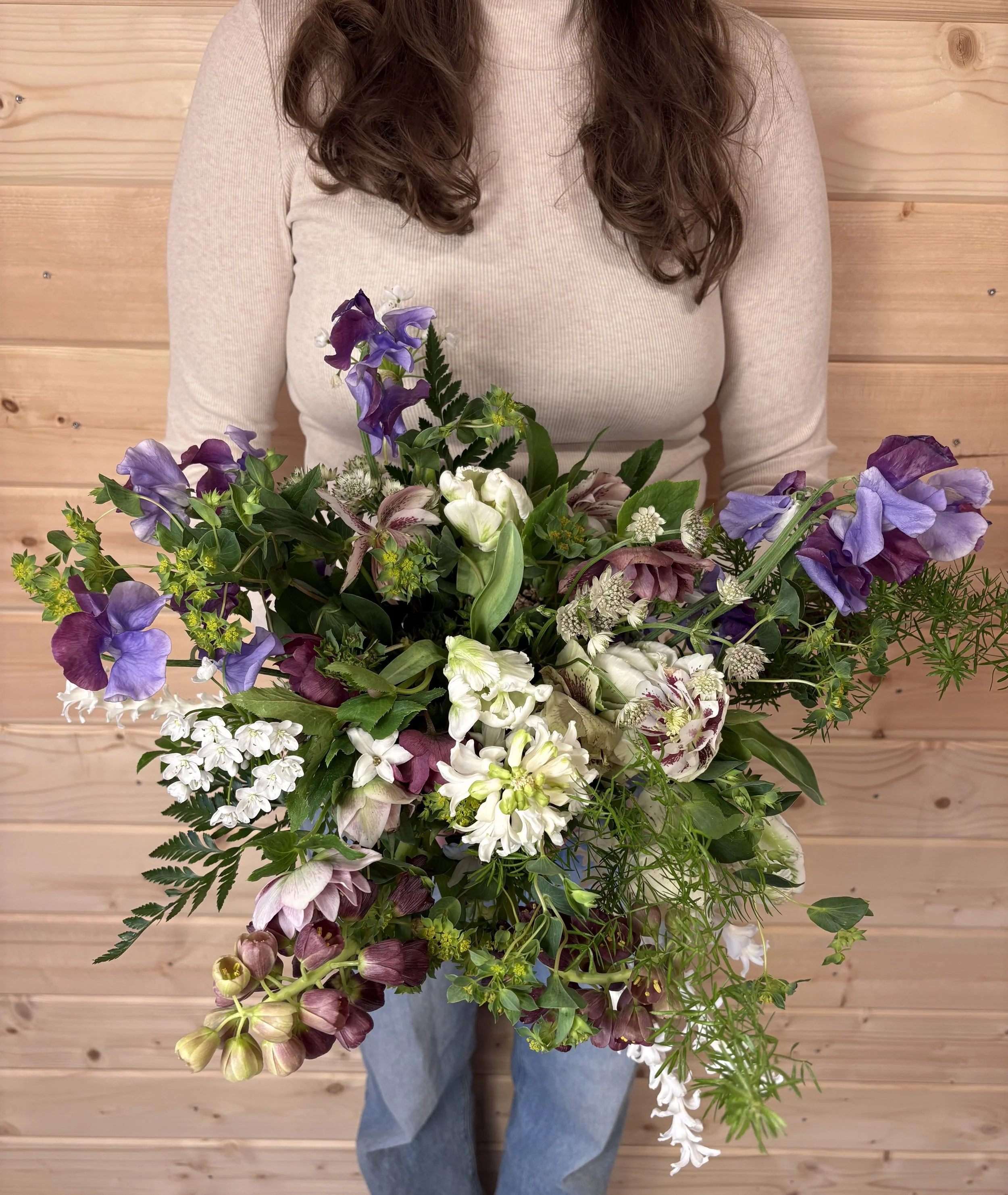 Person holding large bouquet of purple and white flowers in front of a wooden wall.